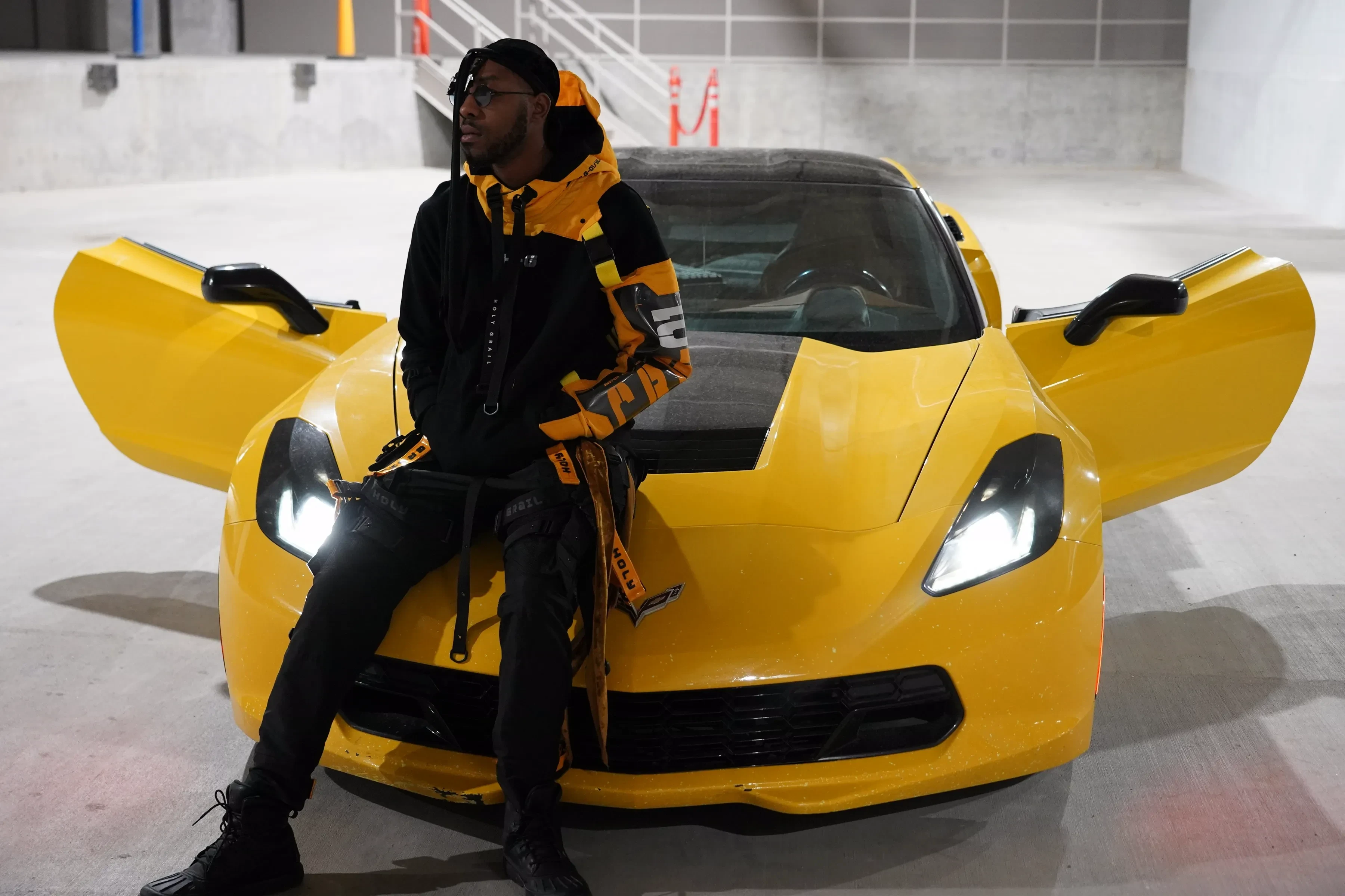 Alpha The Wolf sitting on the front of the Corvette with both doors raised like wings, hood up and sunglasses on, the headlights throwing hard white light across the concrete floor.