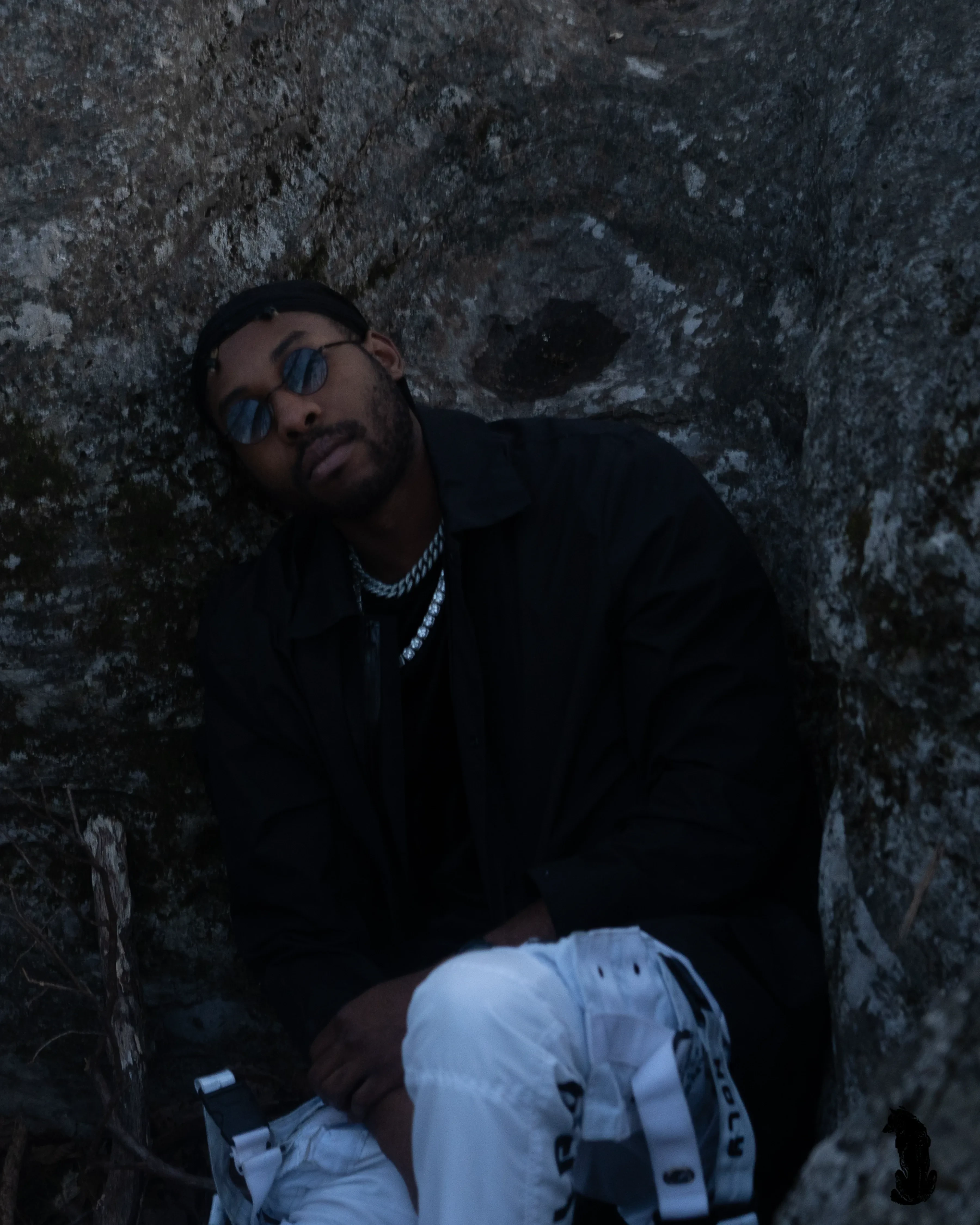 Alpha The Wolf seated in the rock hollow with one hand over his knee and his head propped against the boulder, tinted round glasses and a silver chain in the last of the blue evening light.