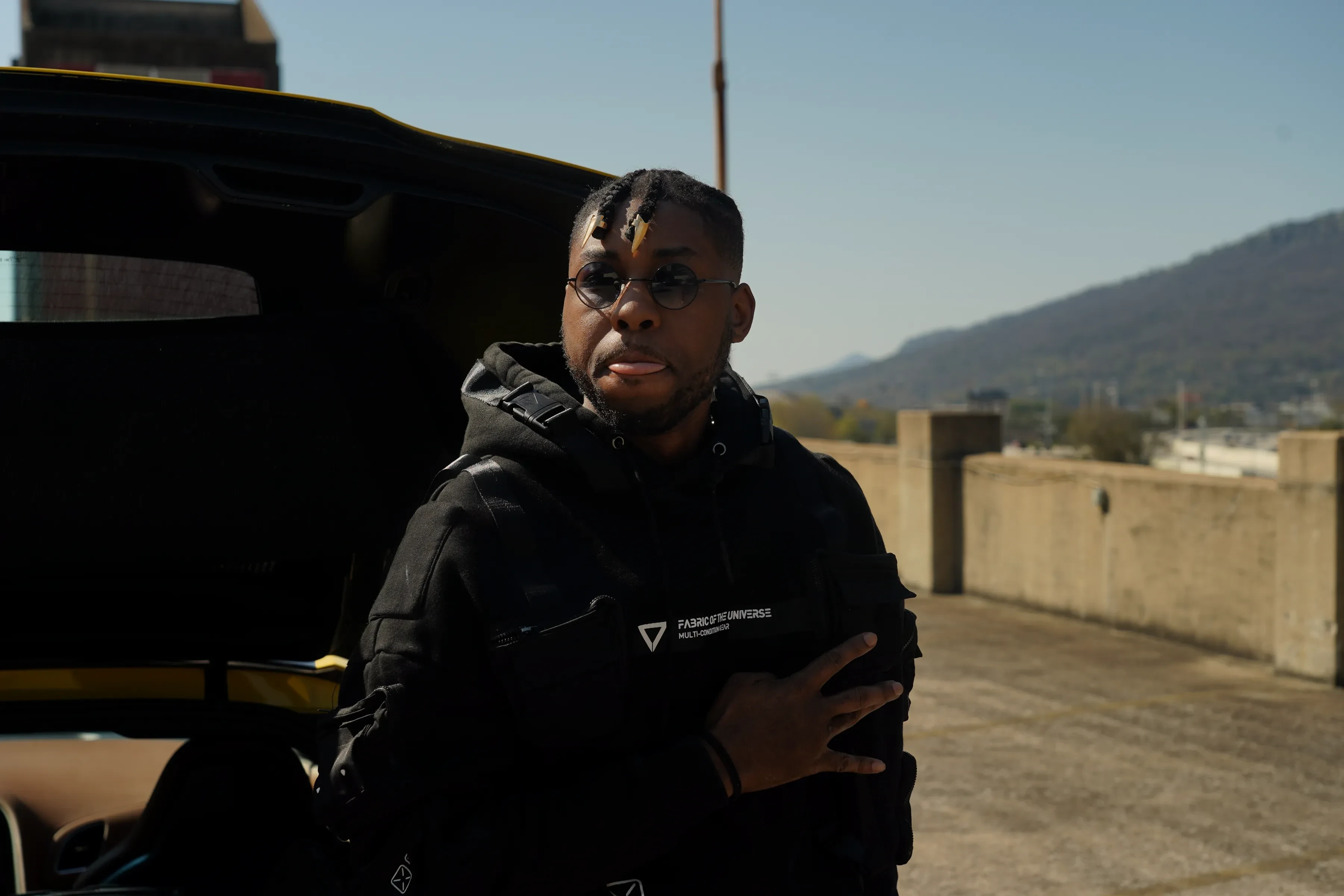 Alpha The Wolf in a close portrait beside the yellow Corvette's raised rear hatch, arms folded across the black jacket, distant hills and a clear sky behind.