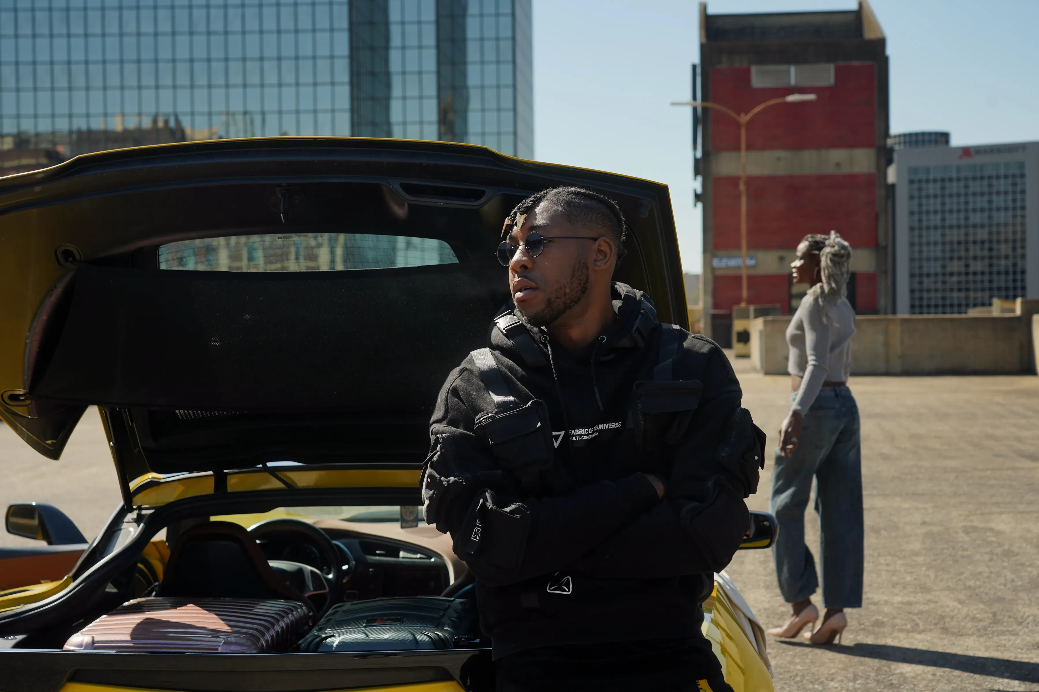 Alpha The Wolf leaning against the Corvette's open hatch with arms crossed and sunglasses on, a person in a grey top walking on the deck behind him, glass towers beyond.