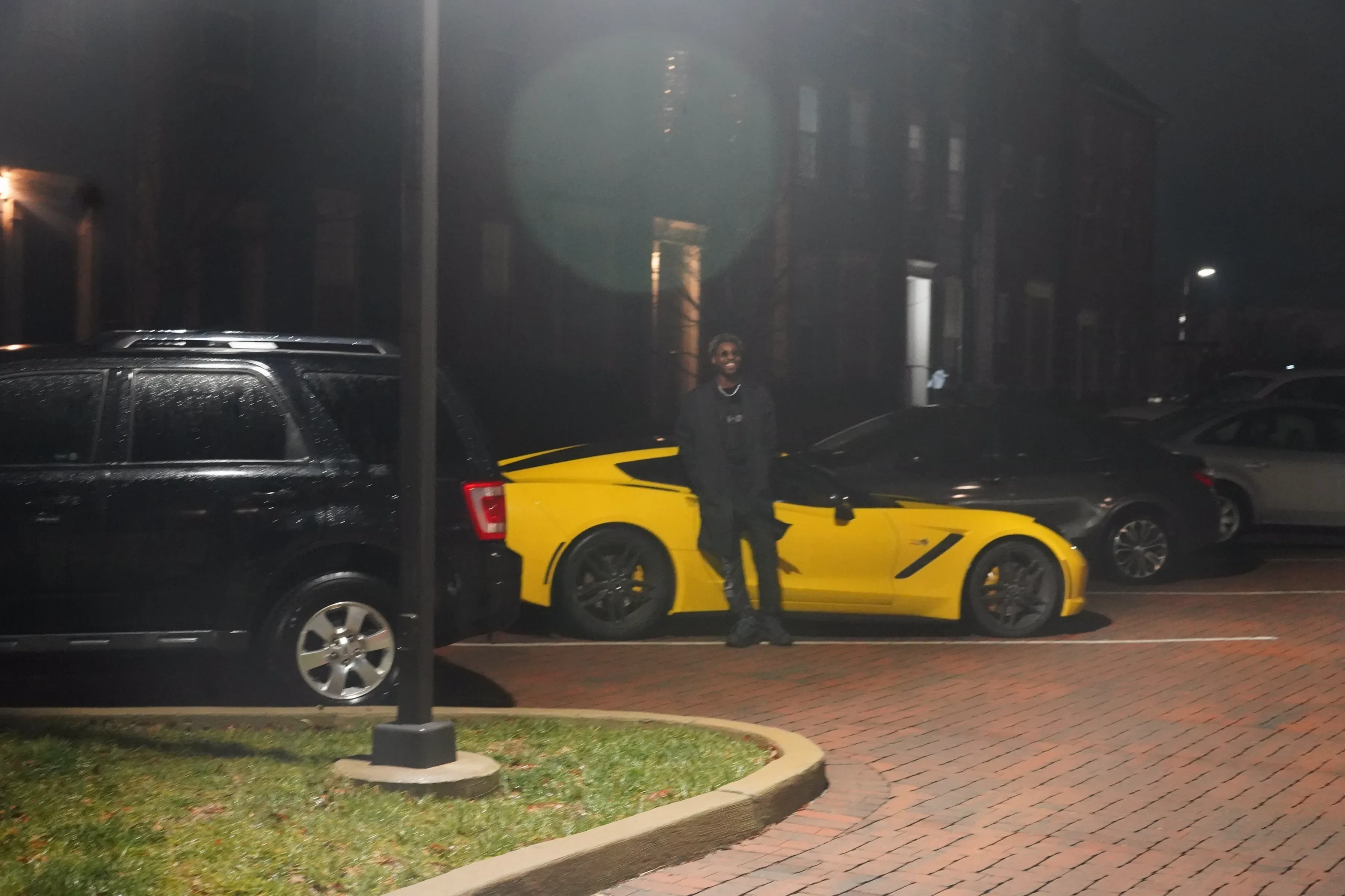 Alpha The Wolf standing between a yellow Corvette and a dark SUV in a misty parking court at night, hands at his sides, streetlamps and townhouse windows glowing faintly through the haze