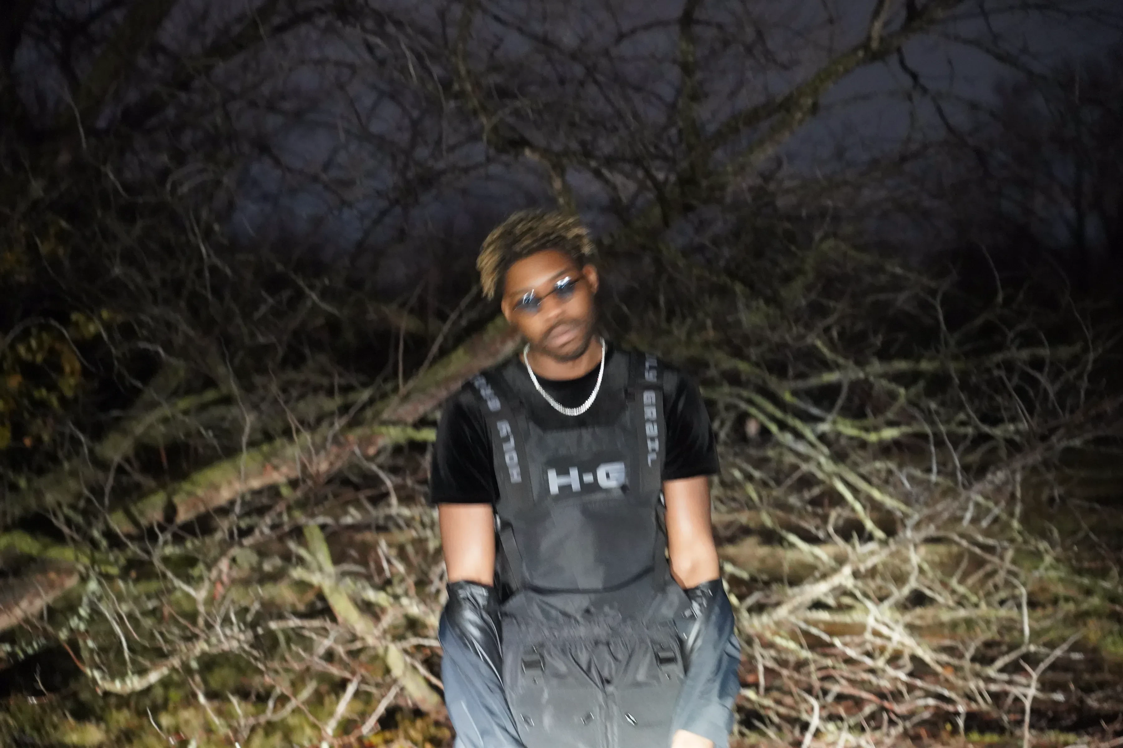 Alpha The Wolf standing among fallen branches in a black vest lettered across the chest, gloved hands at his sides, a chain at his throat and bare trees spread out behind him