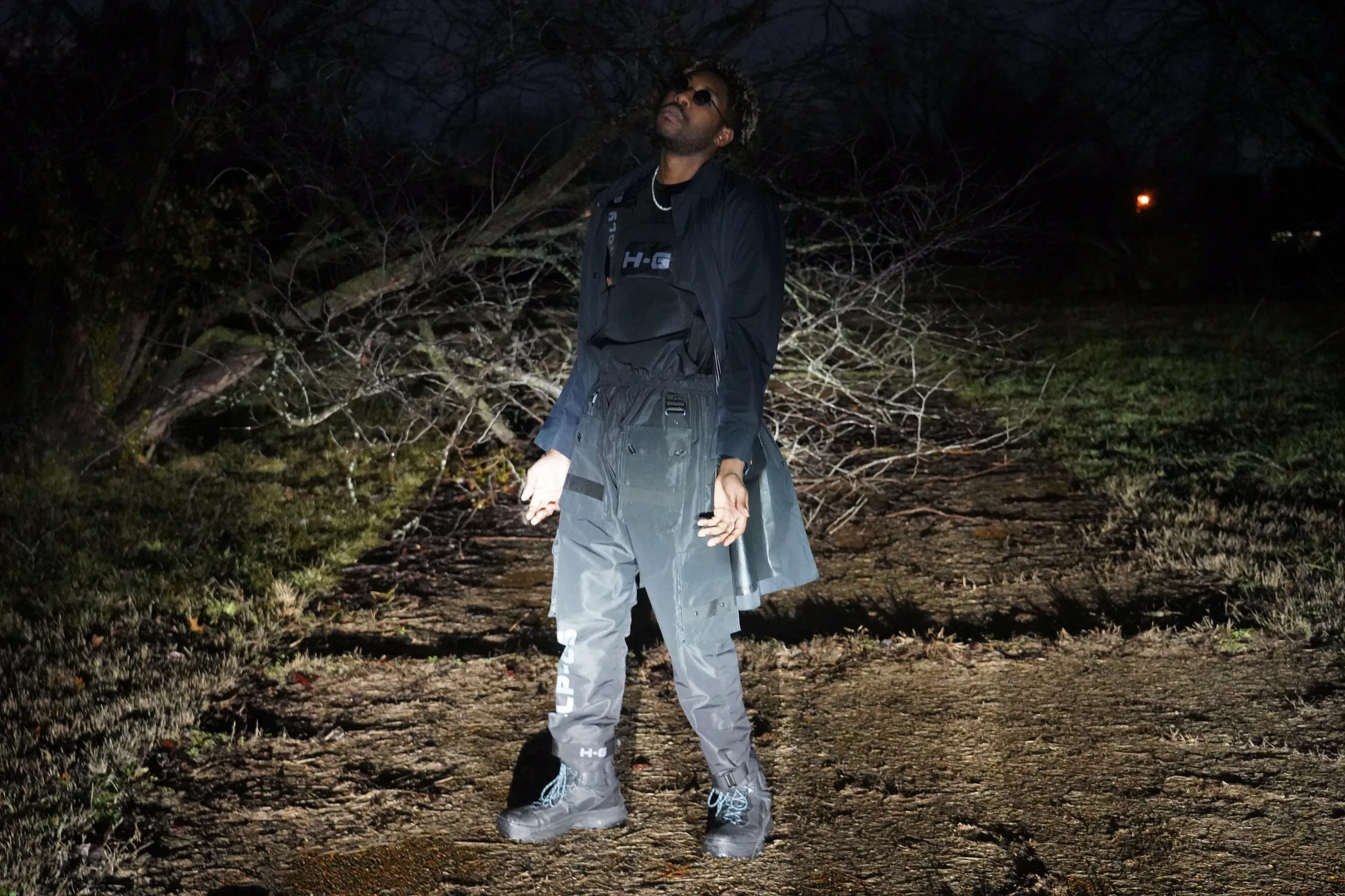 Alpha The Wolf standing full-length in the field with his head tipped back to the sky, jacket open over the vest and cargo pants, bare trees and dark grass around him