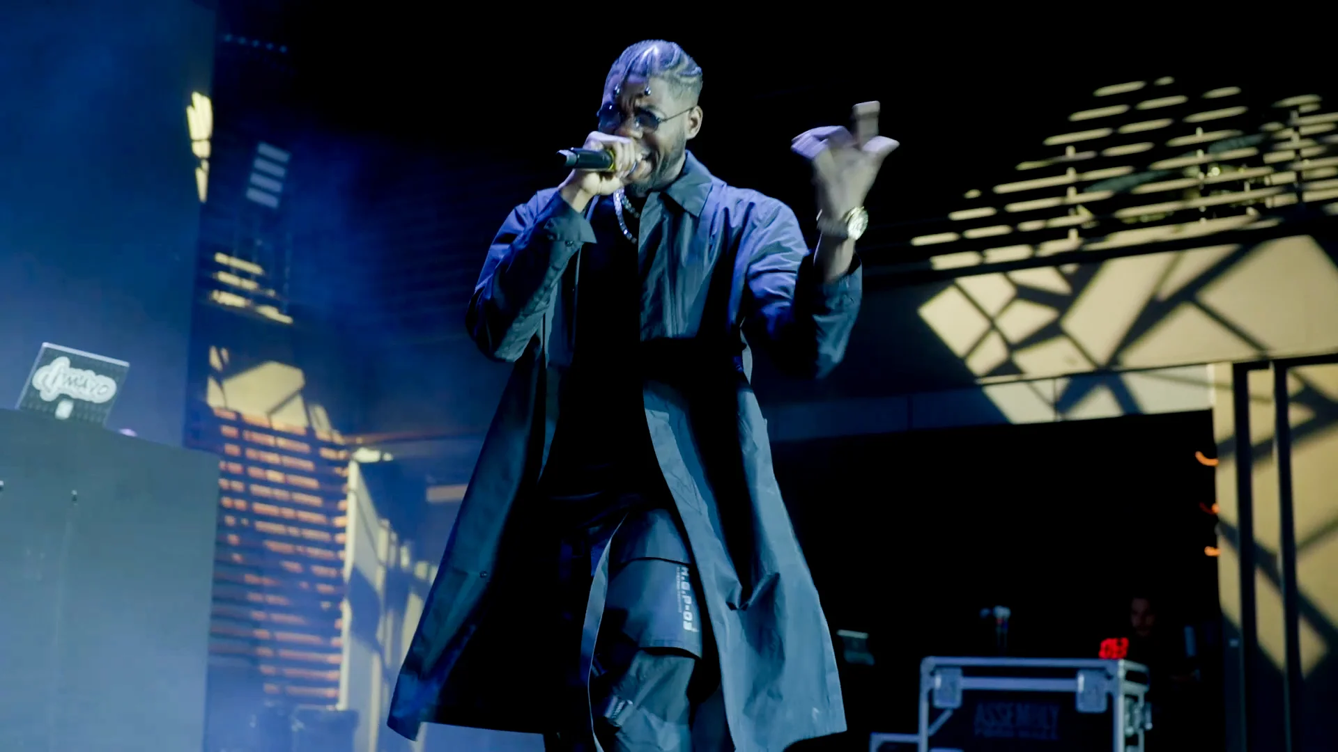 Alpha The Wolf performing on a large stage, mic raised to his mouth and one hand gesturing outward, draped in a long dark trench coat over all-black, bathed in cool blue stage lighting with geometric shadow patterns cast across the backdrop behind him