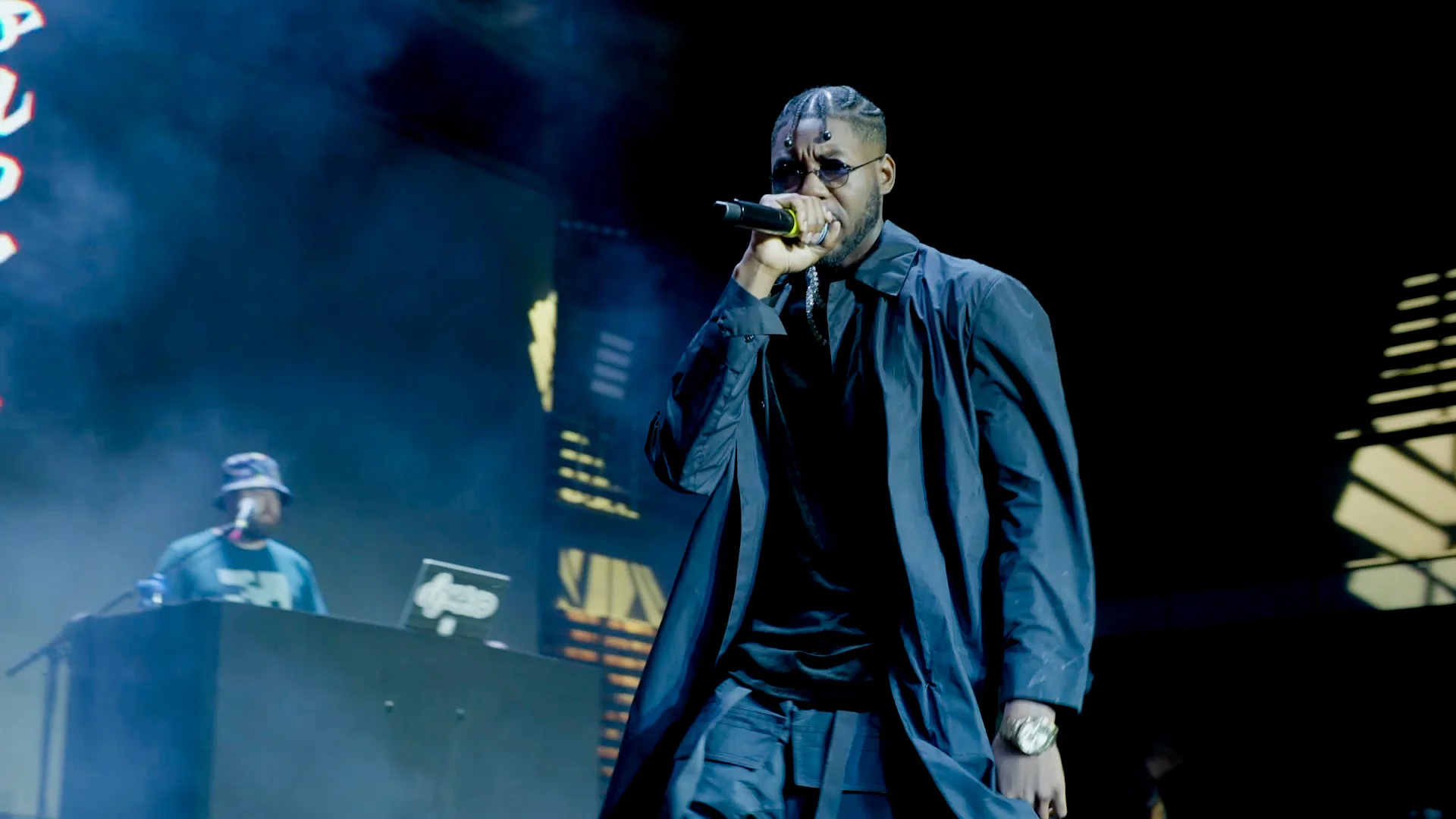 Alpha The Wolf delivering a verse center stage, head slightly bowed over the mic, wearing a long dark overcoat and silver watch, surrounded by rising smoke and deep blue light, with a DJ visible at the decks in the background