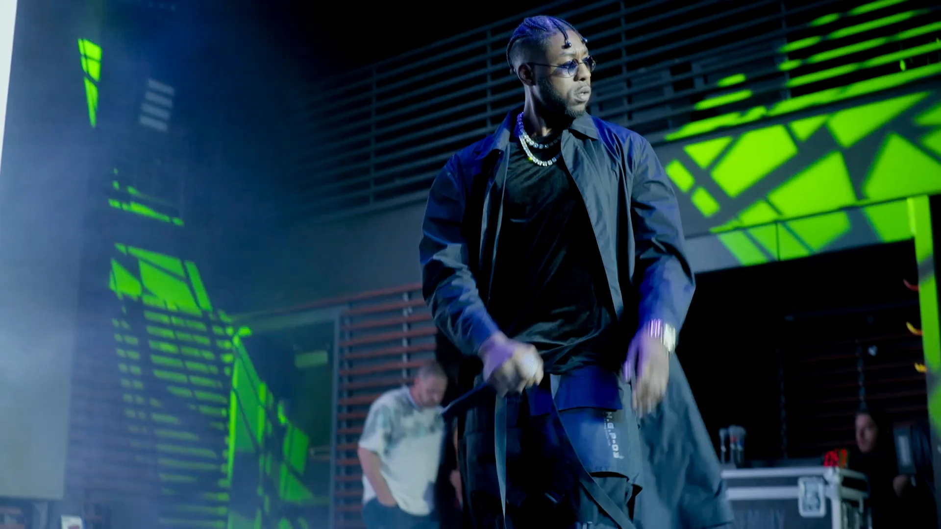 Alpha The Wolf in a commanding stance at the front of the stage, not holding a mic, arms loose and body in motion, wearing a long dark coat and pearl chain, bathed in vivid neon green light that fractures across the stage set behind him