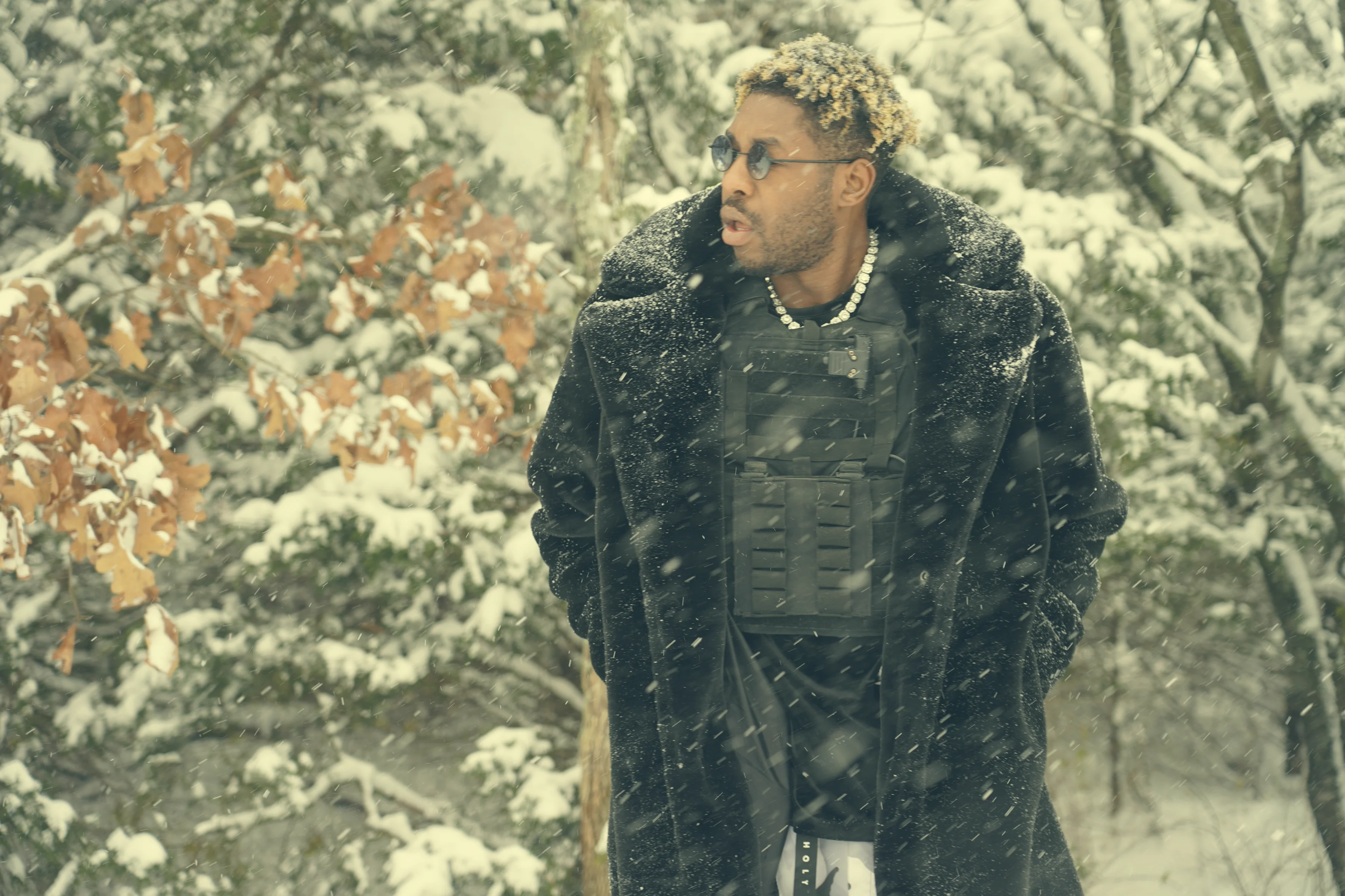 Alpha The Wolf standing in a snow-dusted wooded area, gazing off to one side with a guarded expression, wearing a long black fur-collar coat layered over a tactical vest, pearl chain around his neck, snow falling heavily through the bare trees behind him