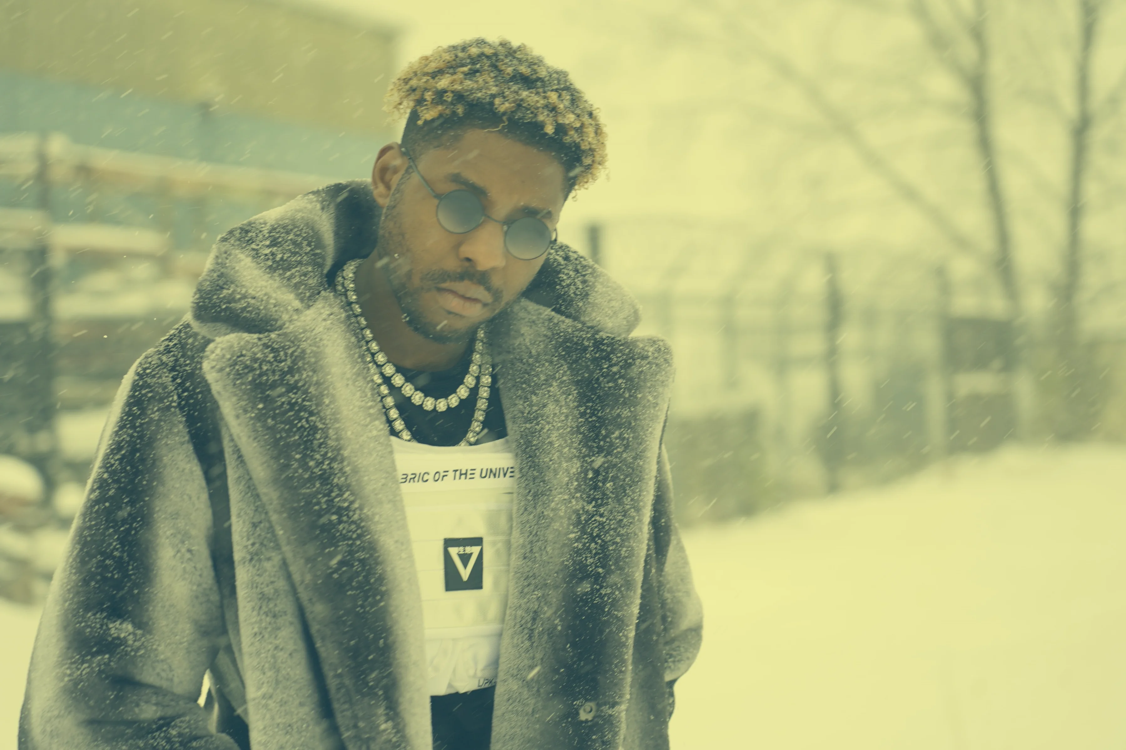 Alpha The Wolf in a closer frame, chin slightly dipped, wearing the same grey shearling coat and pearl chain with round reflective glasses, the yellow-green film tone deepening the winter isolation of the shot as snow continues to fall around him