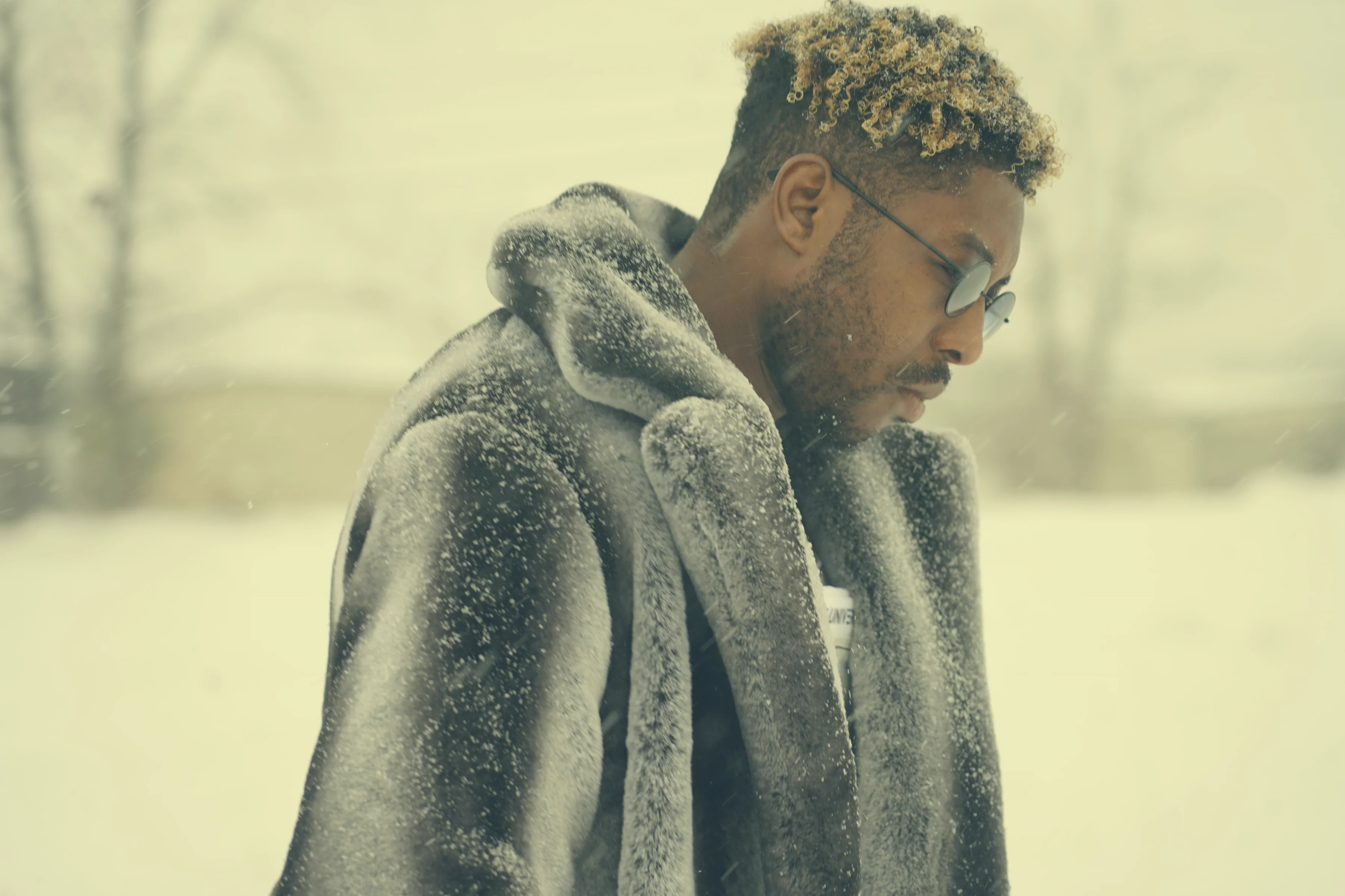 Alpha The Wolf in near-identical profile to the previous frame, head low, expression heavy, the grey shearling coat and round frames unchanged, the blizzard rendering the background a near-white void