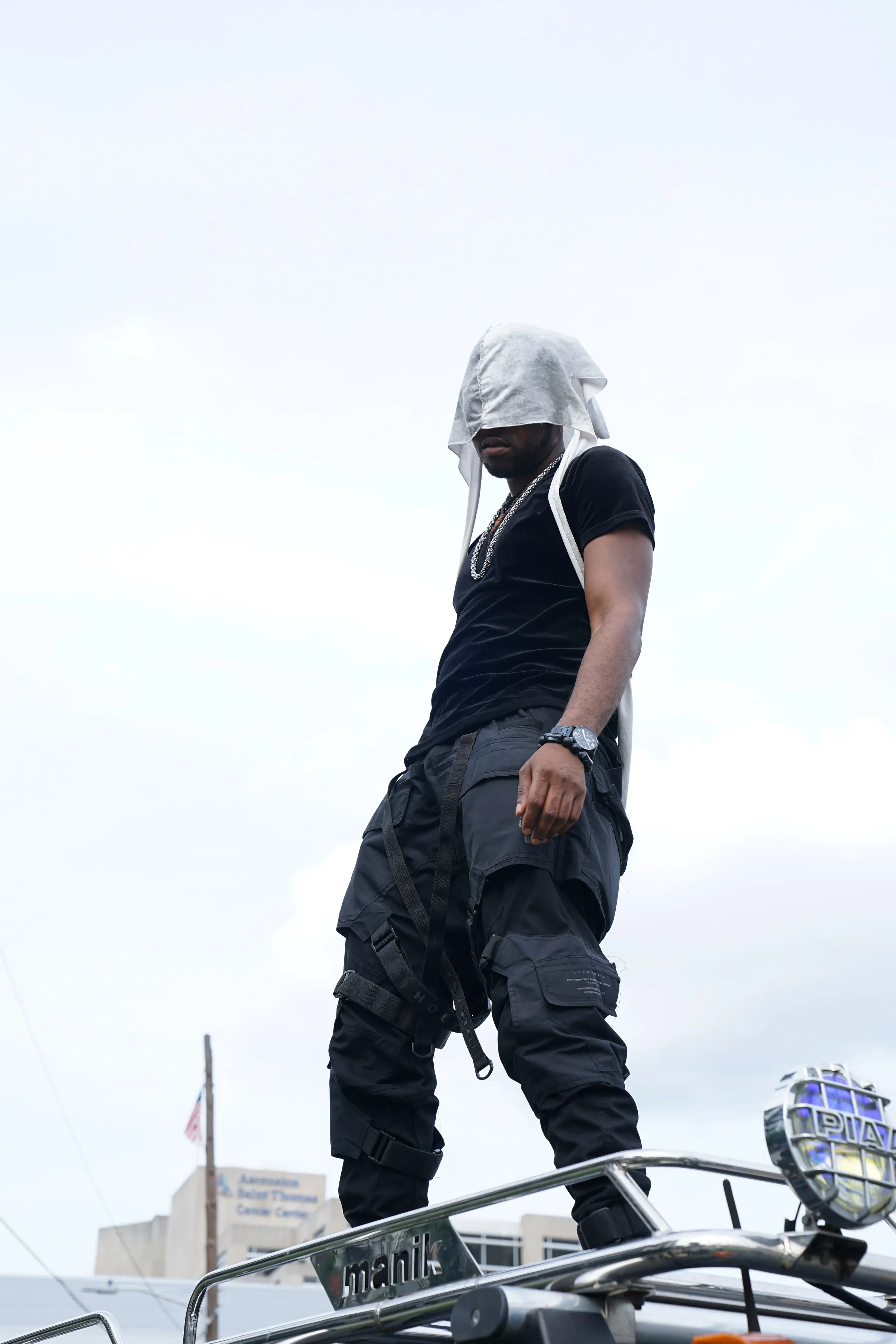 Alpha The Wolf standing on the chrome roof rack of the black Hummer against a flat white Nashville sky, head bowed under a white cloth head-wrap, black velvet tee and black tactical cargo pants