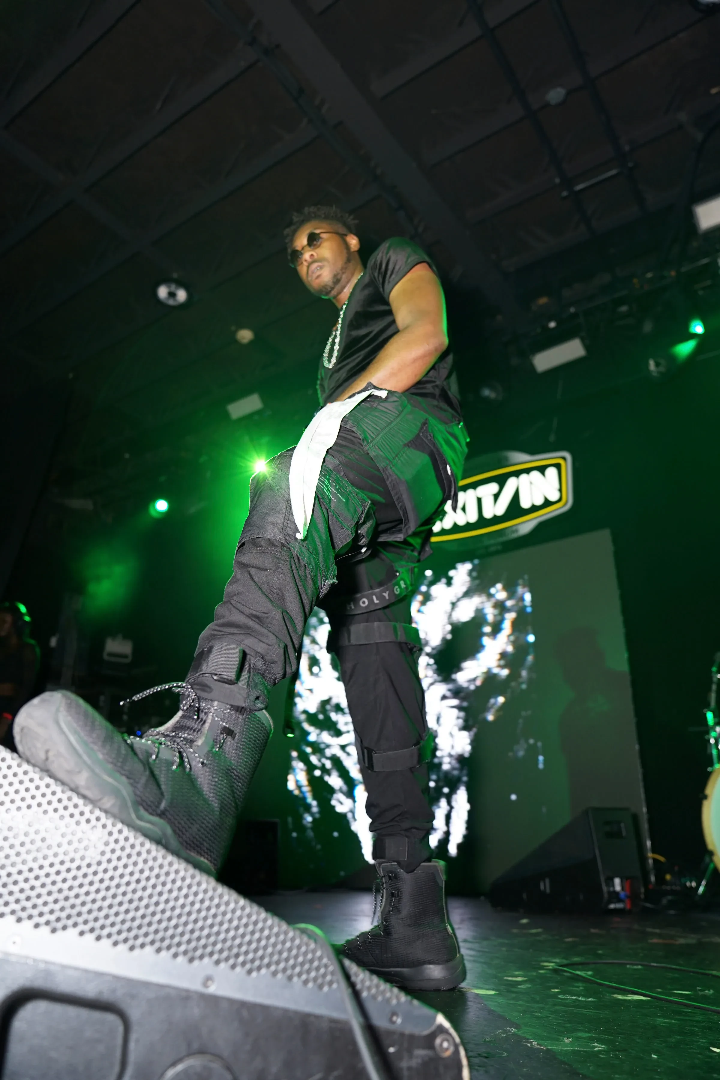 Alpha The Wolf lunging across the Exit/In stage in a low-angle frame lit vivid green, one leg lifted onto a monitor wedge, black velvet tee, strapped cargo pants and black boots, the yellow Exit/In logo glowing on the backdrop