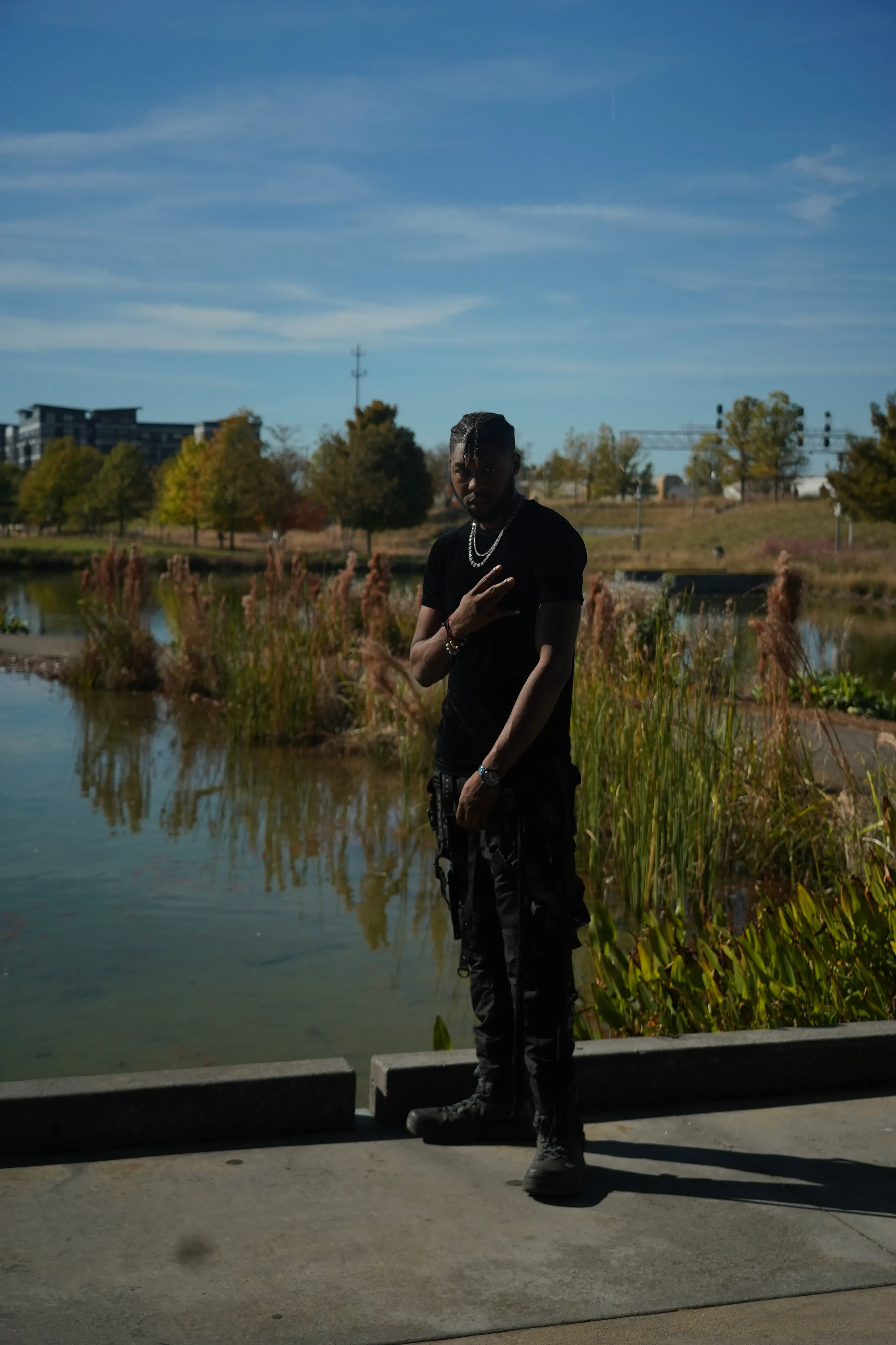 Alpha The Wolf at the concrete edge of a park pond with one hand laid flat on his chest, dressed head to toe in black with a silver cuban chain, rust-coloured reeds and an apartment block across the water behind him