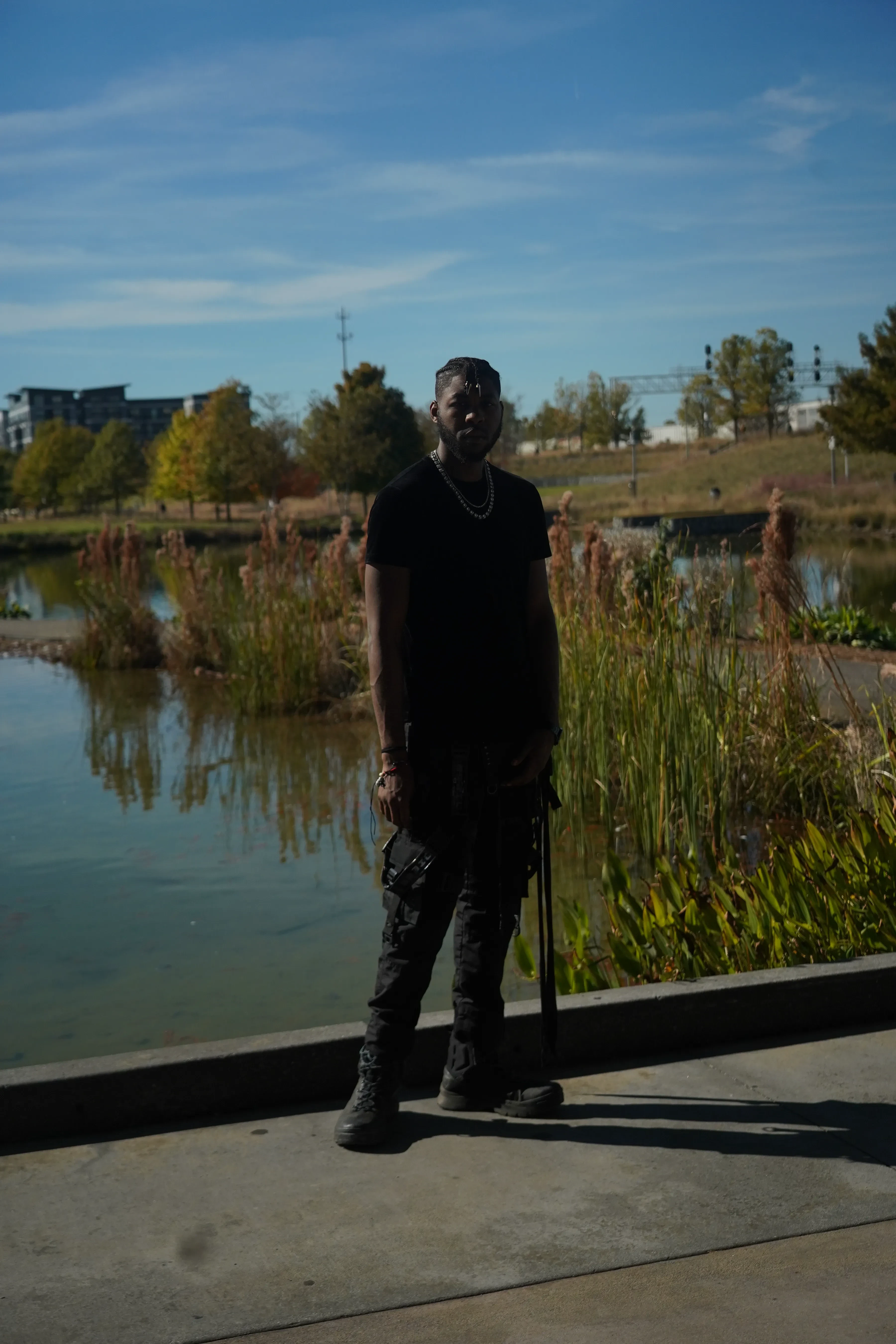 Alpha The Wolf standing square to the camera at the water's edge in full black, a silver chain at his collar and a beaded bracelet on one wrist, his long shadow thrown across the pale concrete