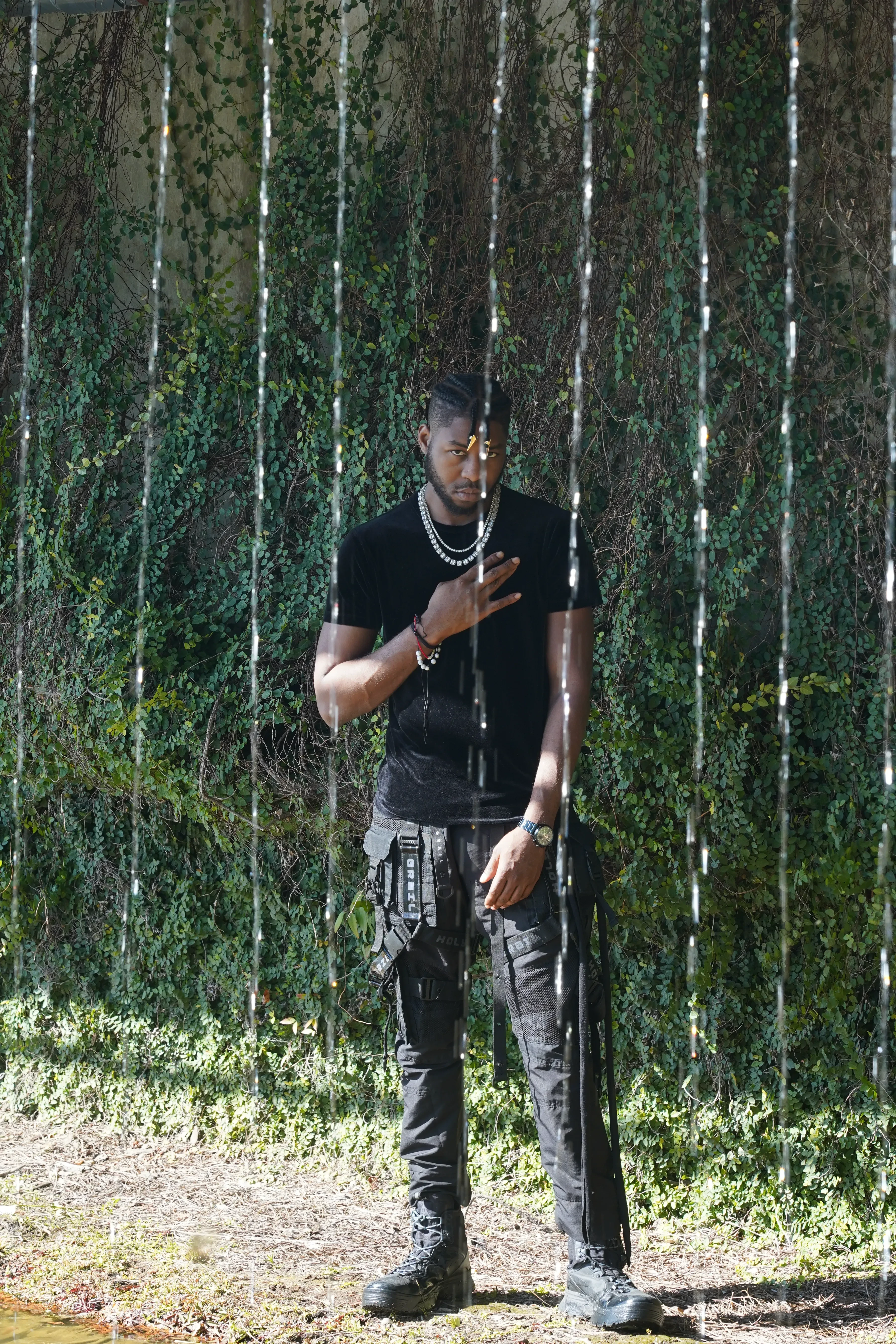 Alpha The Wolf standing beneath the bridge with one arm raised in front of him, black tactical pants with a strapped utility pouch, water streaming down past him on both sides