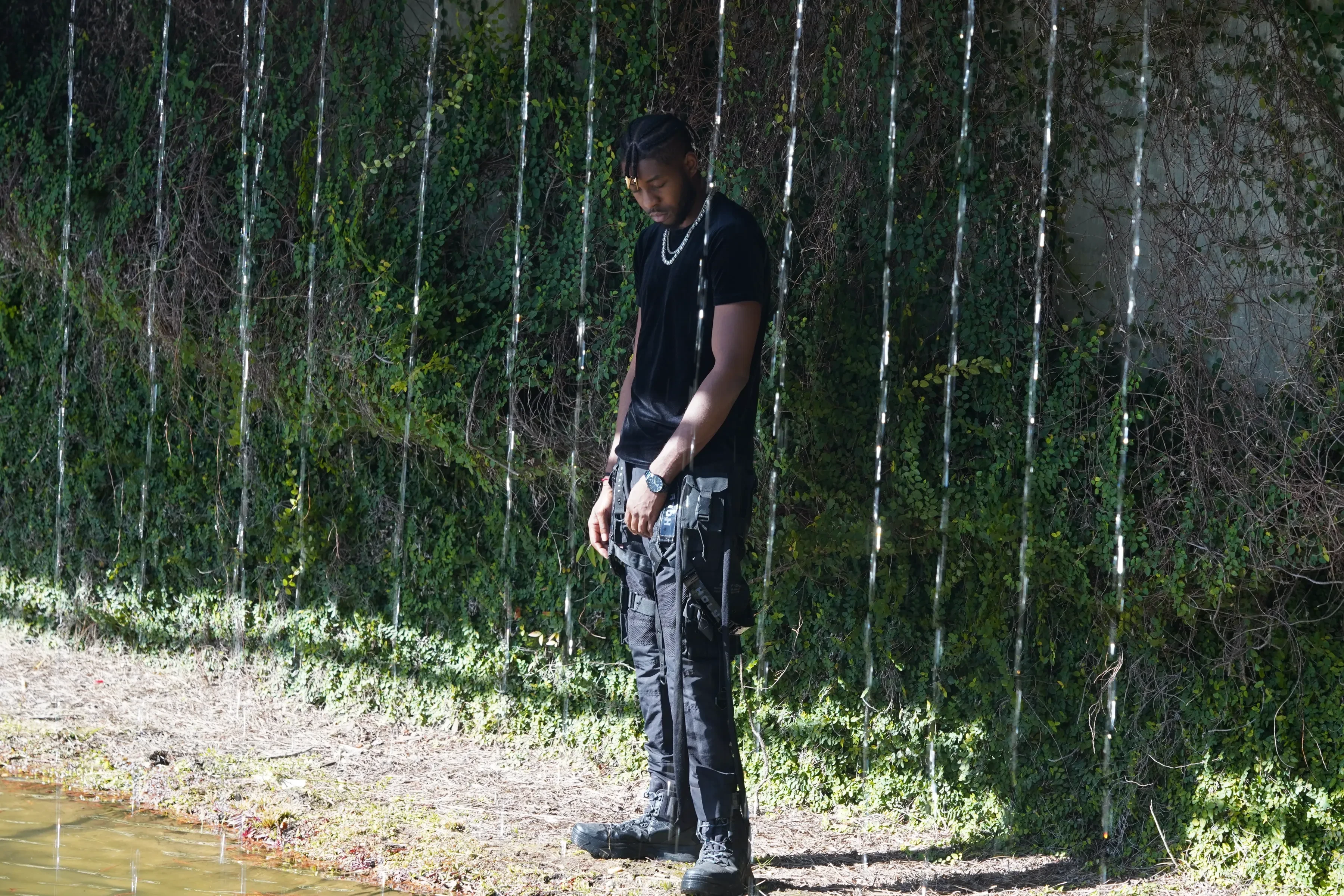 Alpha The Wolf with his head down and hands hanging low, black boots planted on the dry bank, water pouring in long unbroken strands all around him against a wall of dense green ivy