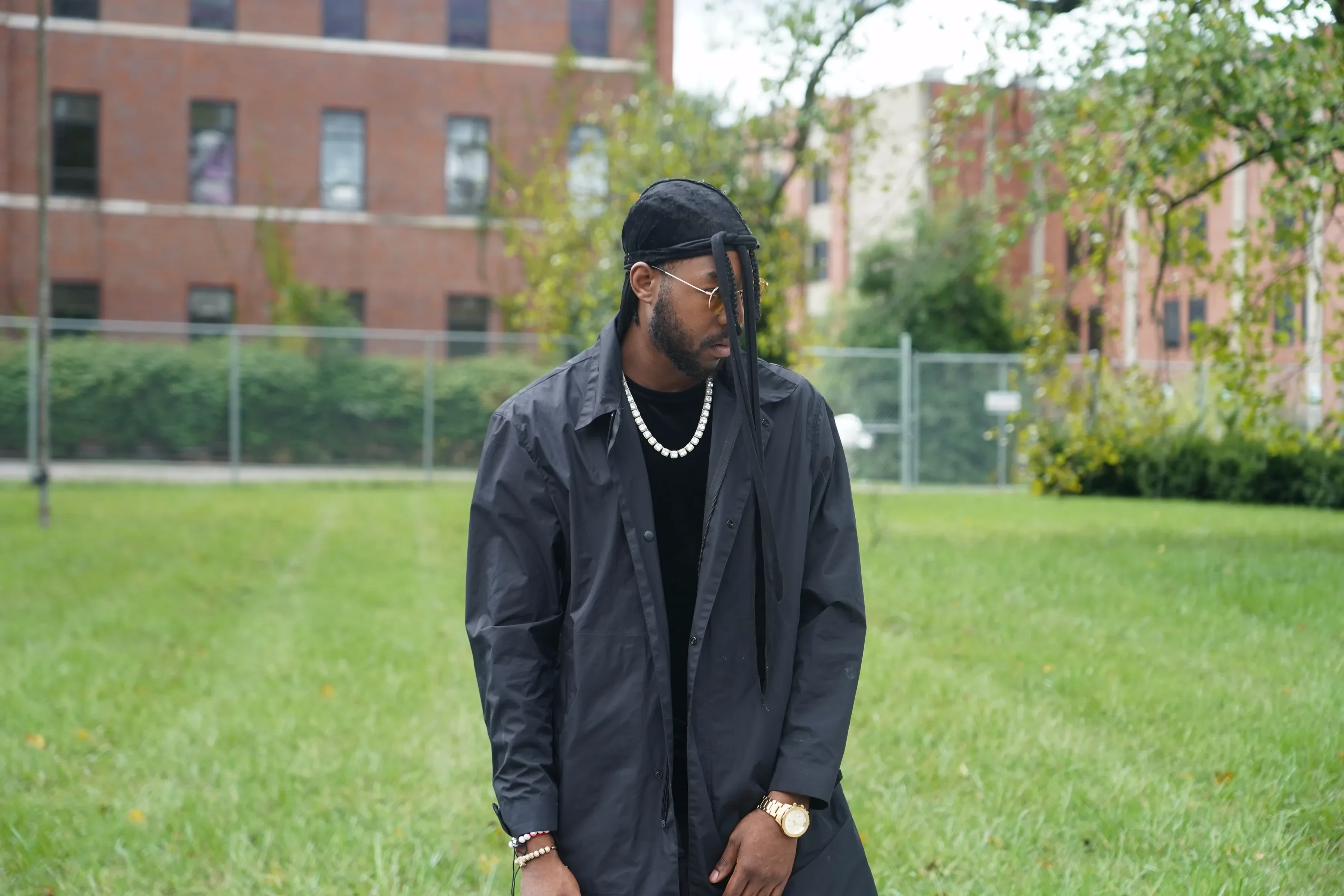 Alpha The Wolf standing on mown grass with his head bowed and both arms hanging loose, a black trench open over a black tee and white bead chain, a red brick building with rows of dark windows behind him