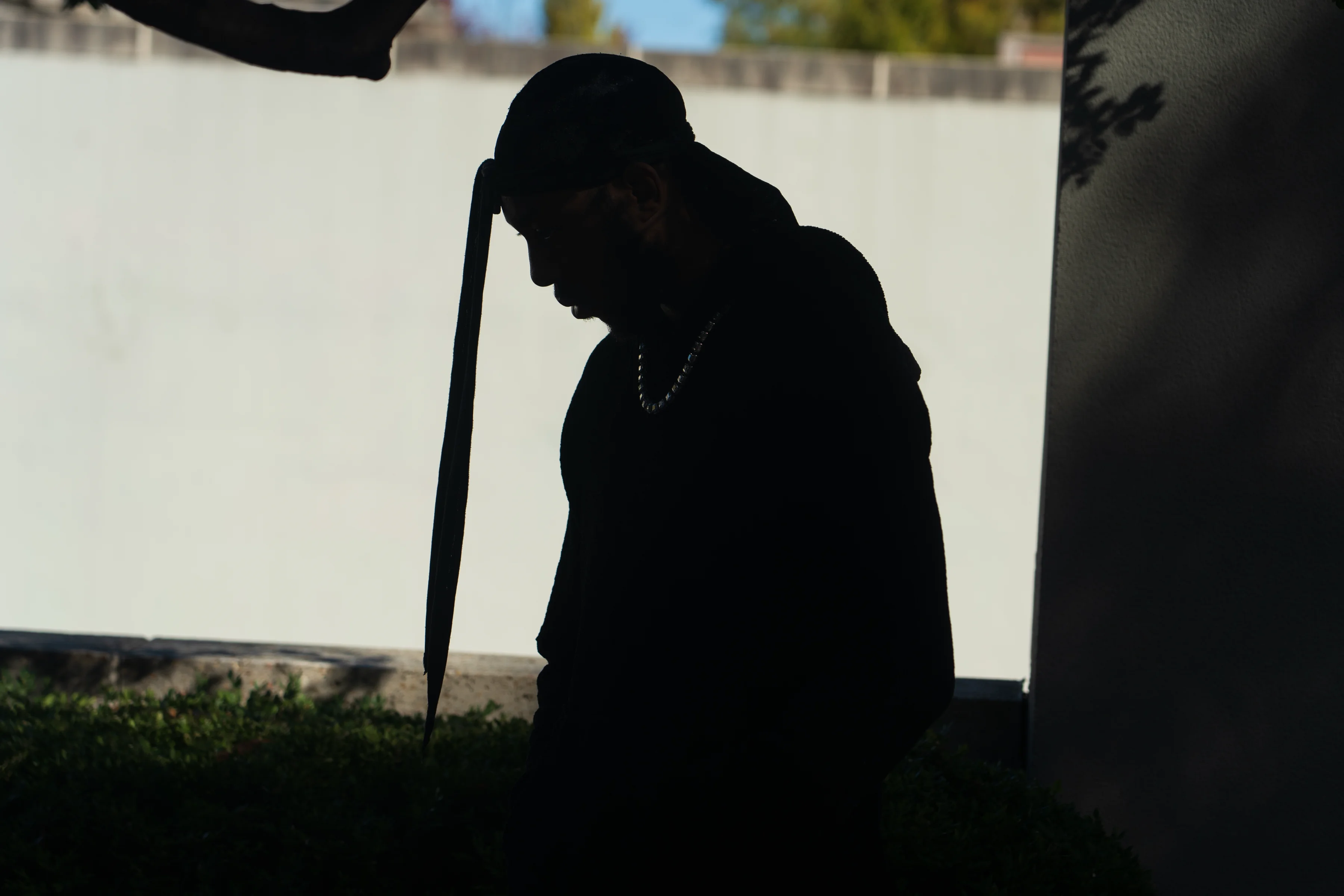 Alpha The Wolf rendered almost entirely in silhouette against a white sunlit wall, a long strap hanging from his cap and a pearl chain glinting at his collar