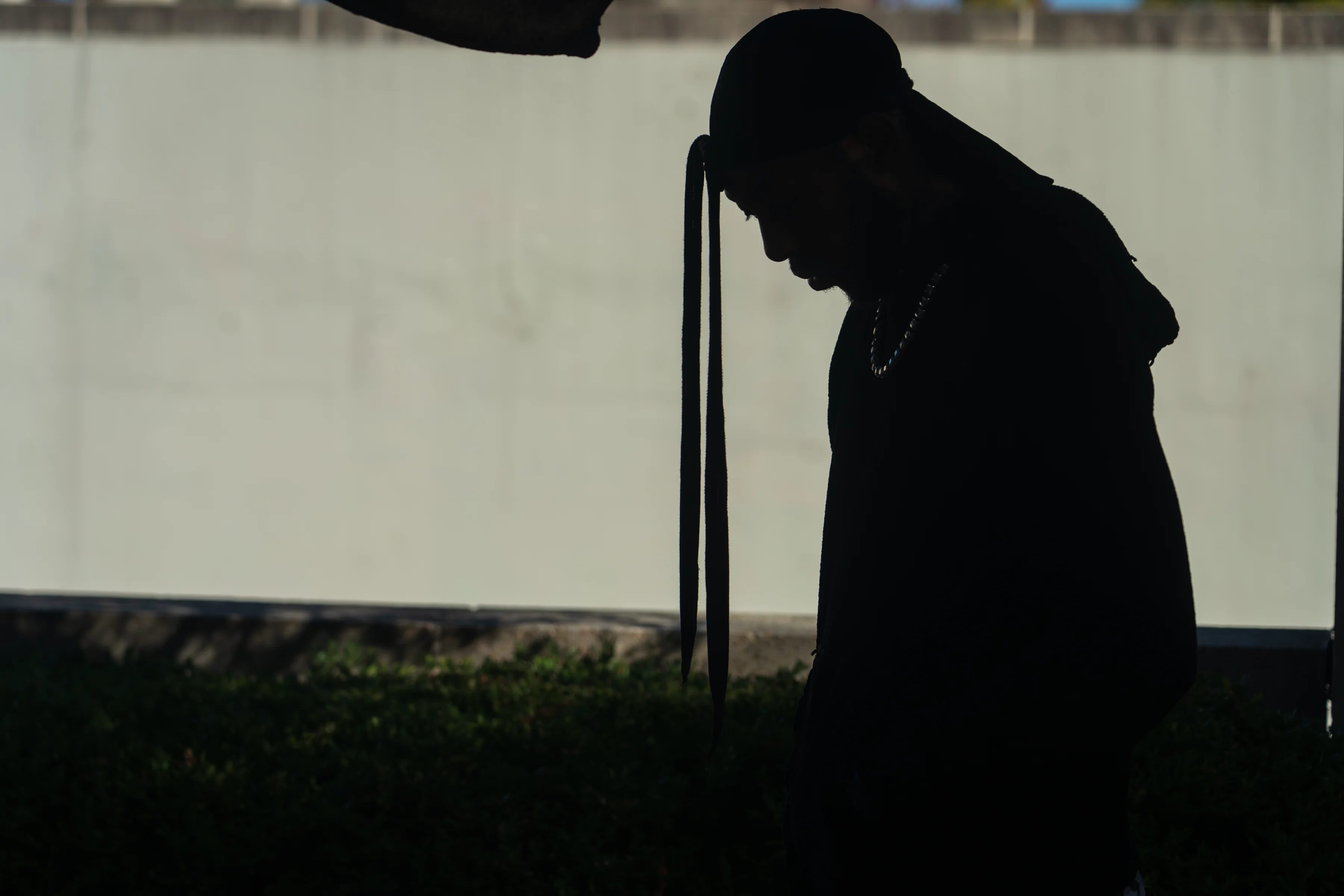 Alpha The Wolf in profile silhouette, head dropped low and a long strap from his cap falling across his face, his turquoise beaded necklace the only colour in the frame