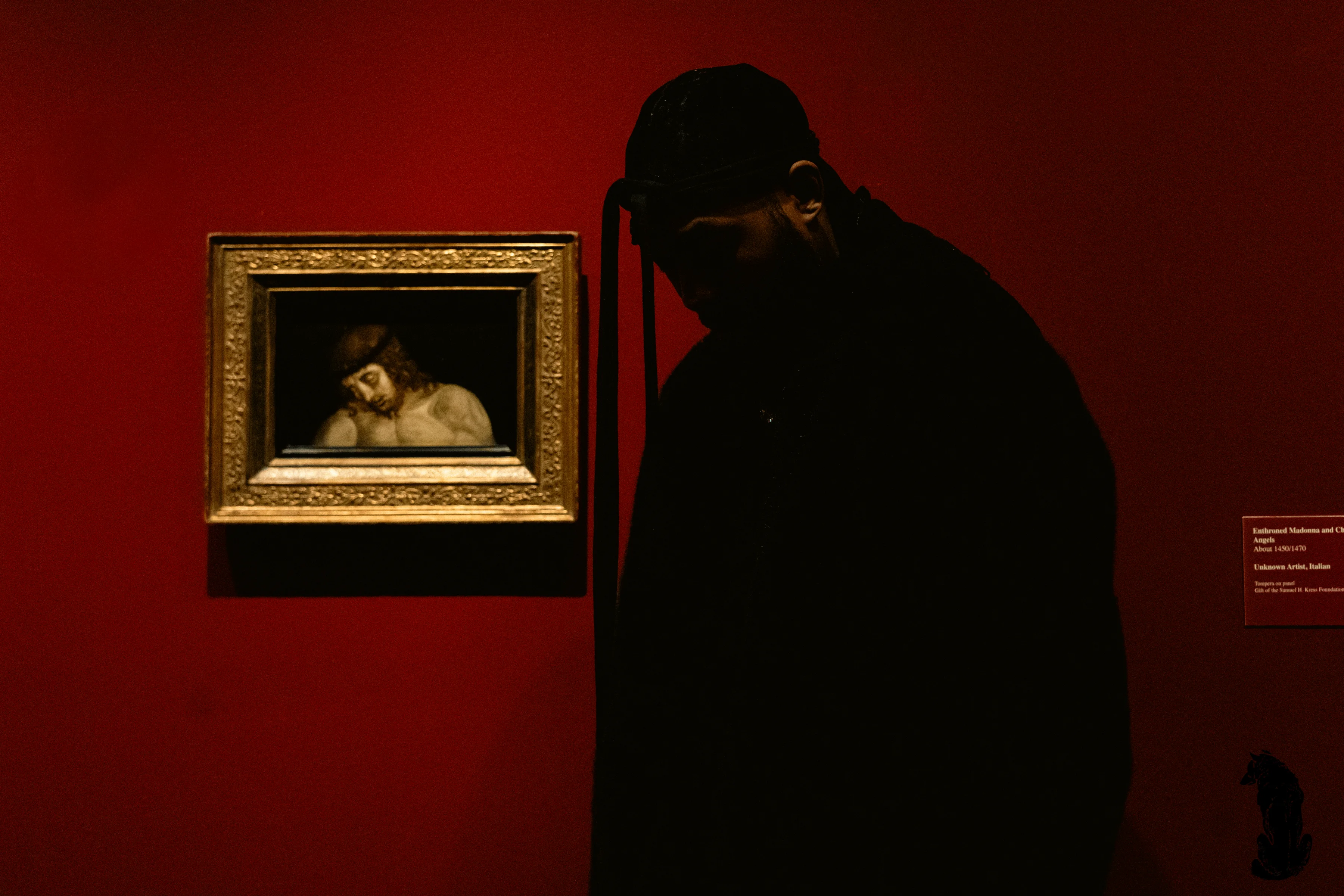 Alpha The Wolf leaning into a small gilded painting on a deep crimson museum wall, his silhouette nearly merging with the darkness as he peers at a classical religious work