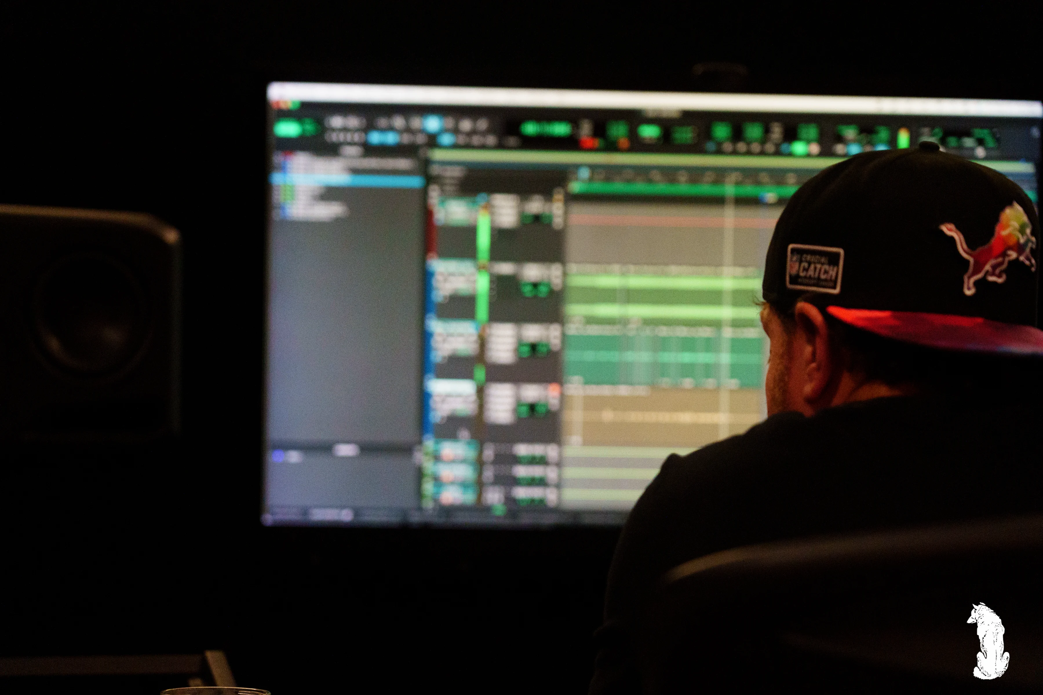 Alpha The Wolf seen from behind in close-up, focused on a brightly lit DAW arrangement screen full of colorful audio tracks, wearing a black tee and a red-brimmed Detroit Lions snapback, the studio speakers flanking the monitor in the darkness