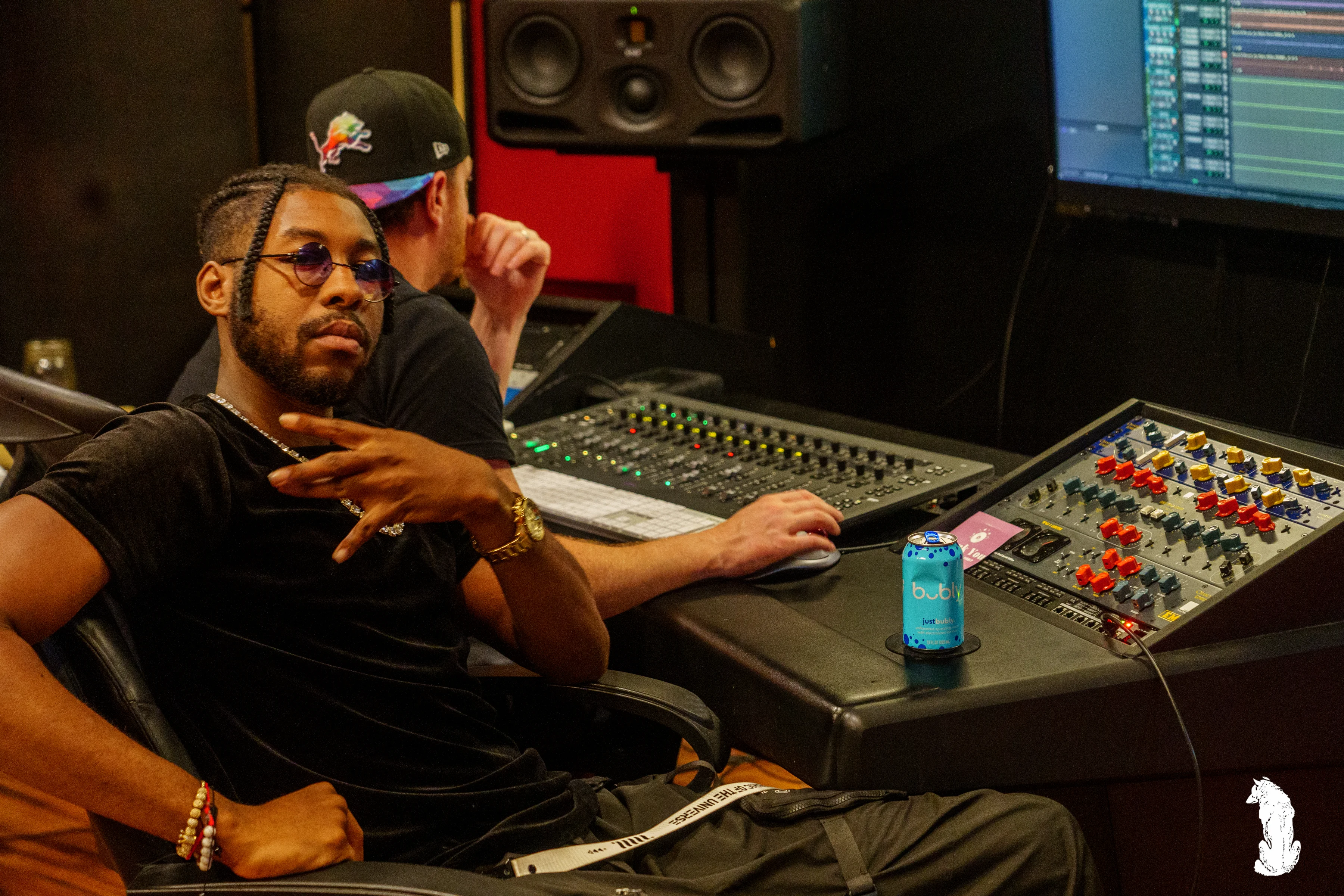 Alpha The Wolf and his producer both leaned in, studying the session intently, Alpha in a black tee and braids with a beaded bracelet and chain, the producer beside him with a colorful snapback, the bright monitor and large-format mixer filling the frame between them