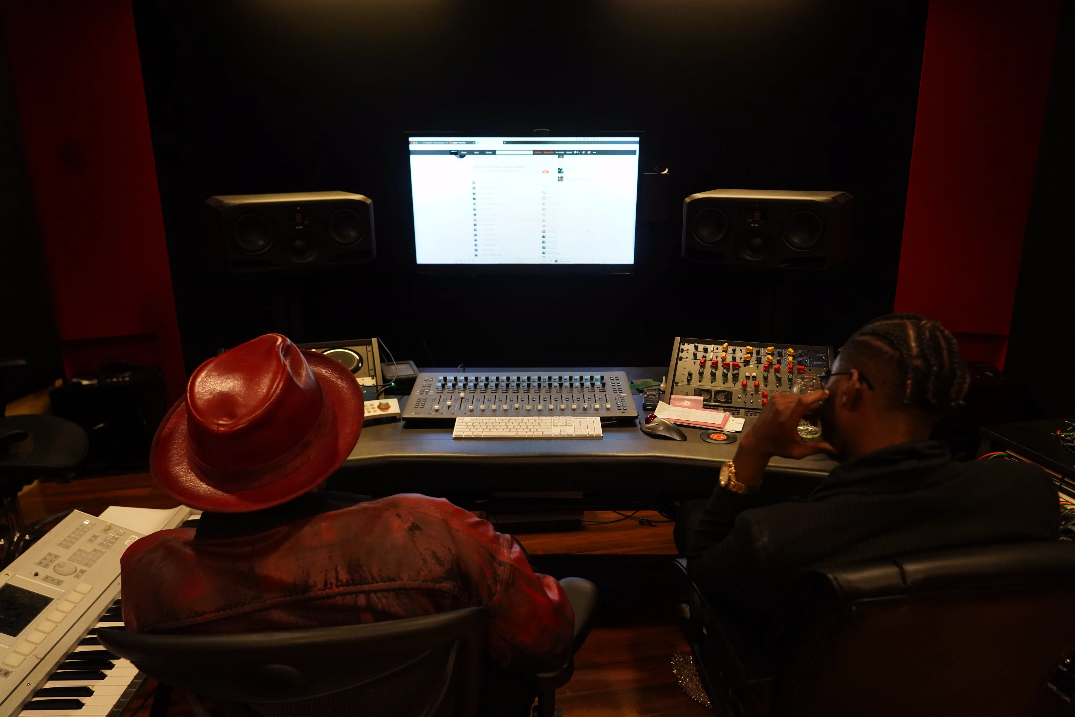 Alpha The Wolf and a collaborator seated at a recording console in a professionally treated studio, both facing a large monitor showing a DAW session, the collaborator wearing a distinctive red wide-brim hat and patterned jacket, a keyboard visible at left