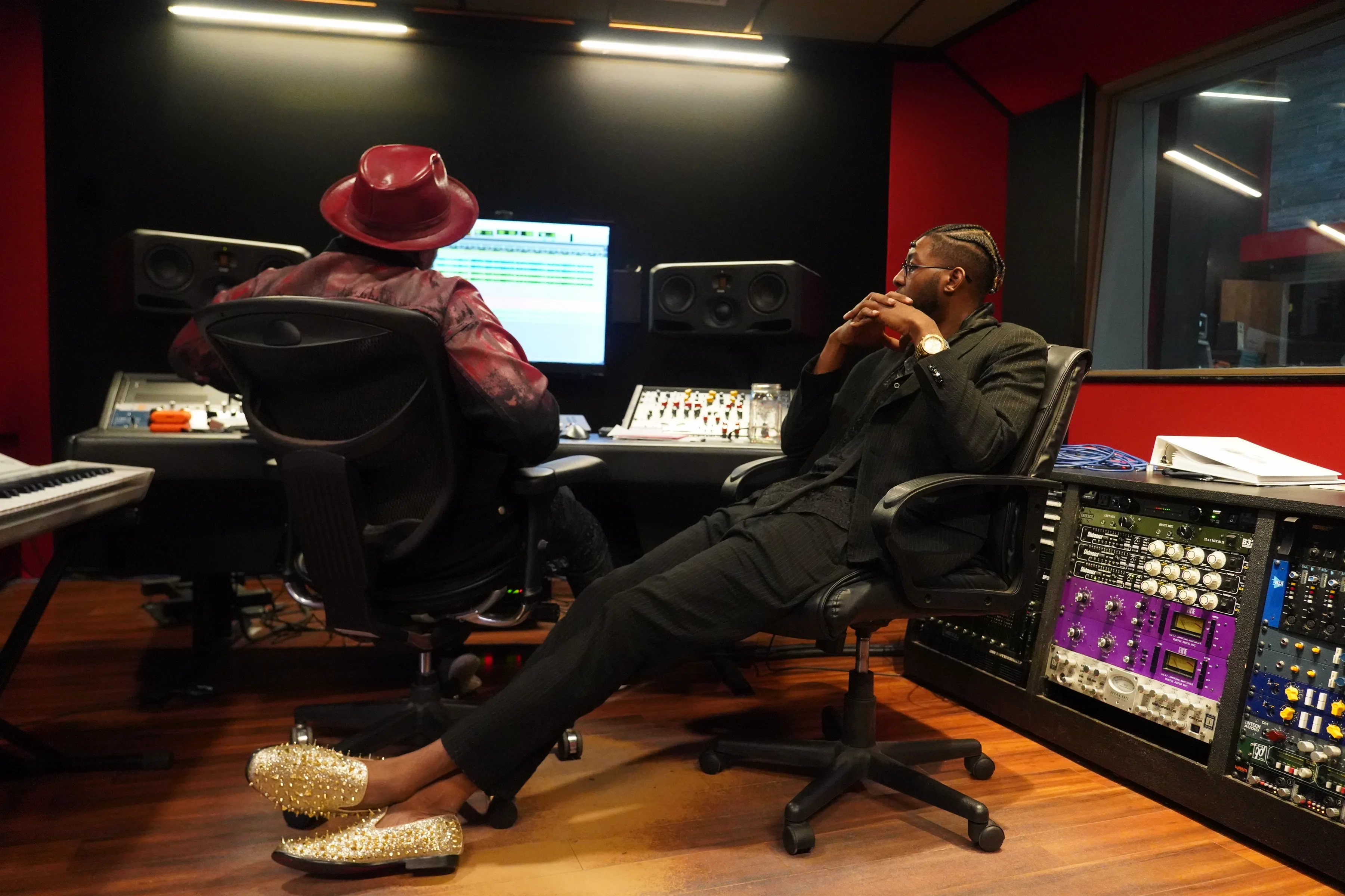 Alpha The Wolf leaned back in a studio chair with legs extended and feet crossed, wearing a black suit and gold loafers with spiked detailing, facing the collaborator seated at the console, the red and black control room decor and rack gear visible behind them