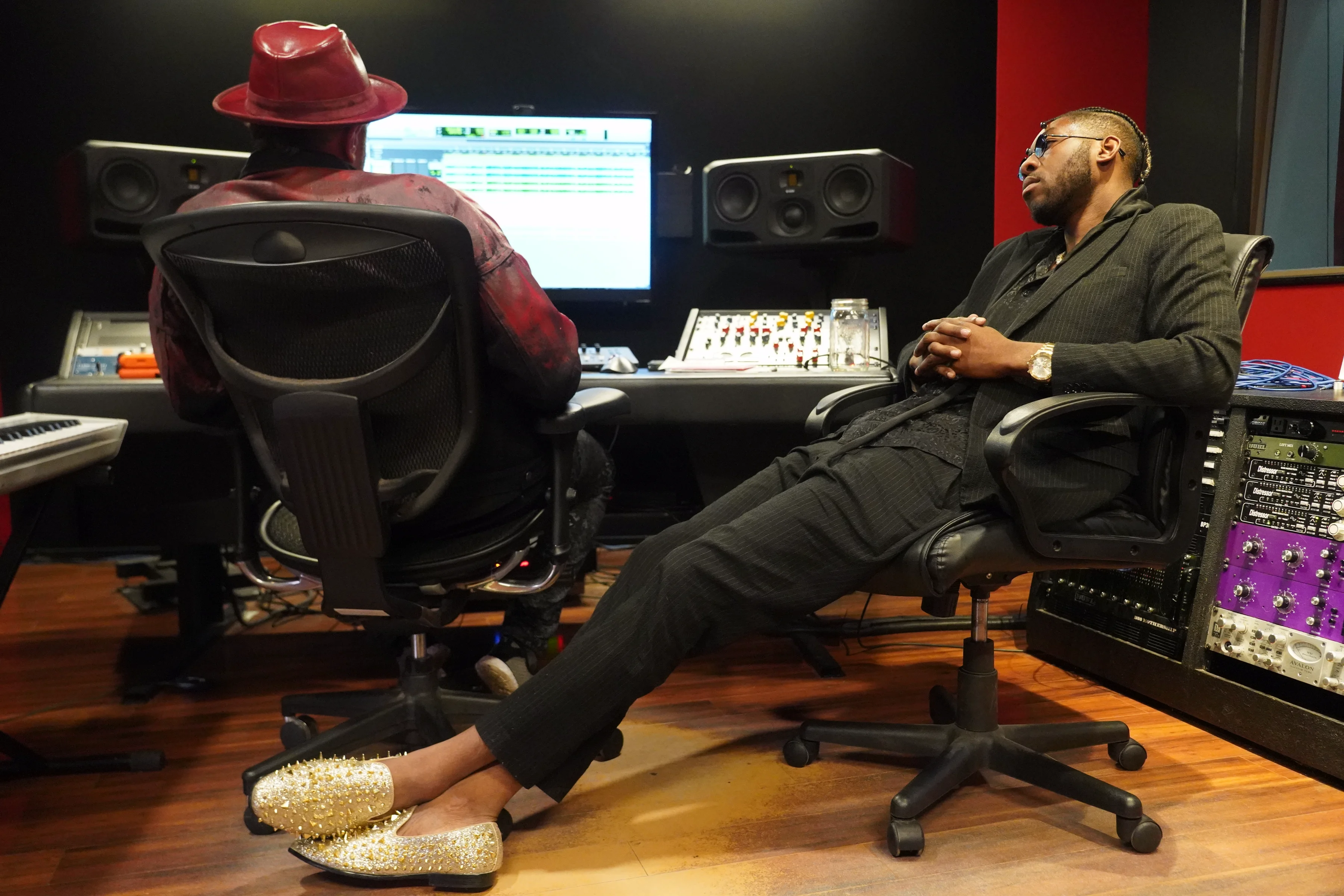 Alpha The Wolf and the collaborator both turned away from the console, deep in back-and-forth dialogue, Alpha in his black suit and gold loafers with arms relaxed, the collaborator in the red hat and leather jacket, the full studio environment visible around them