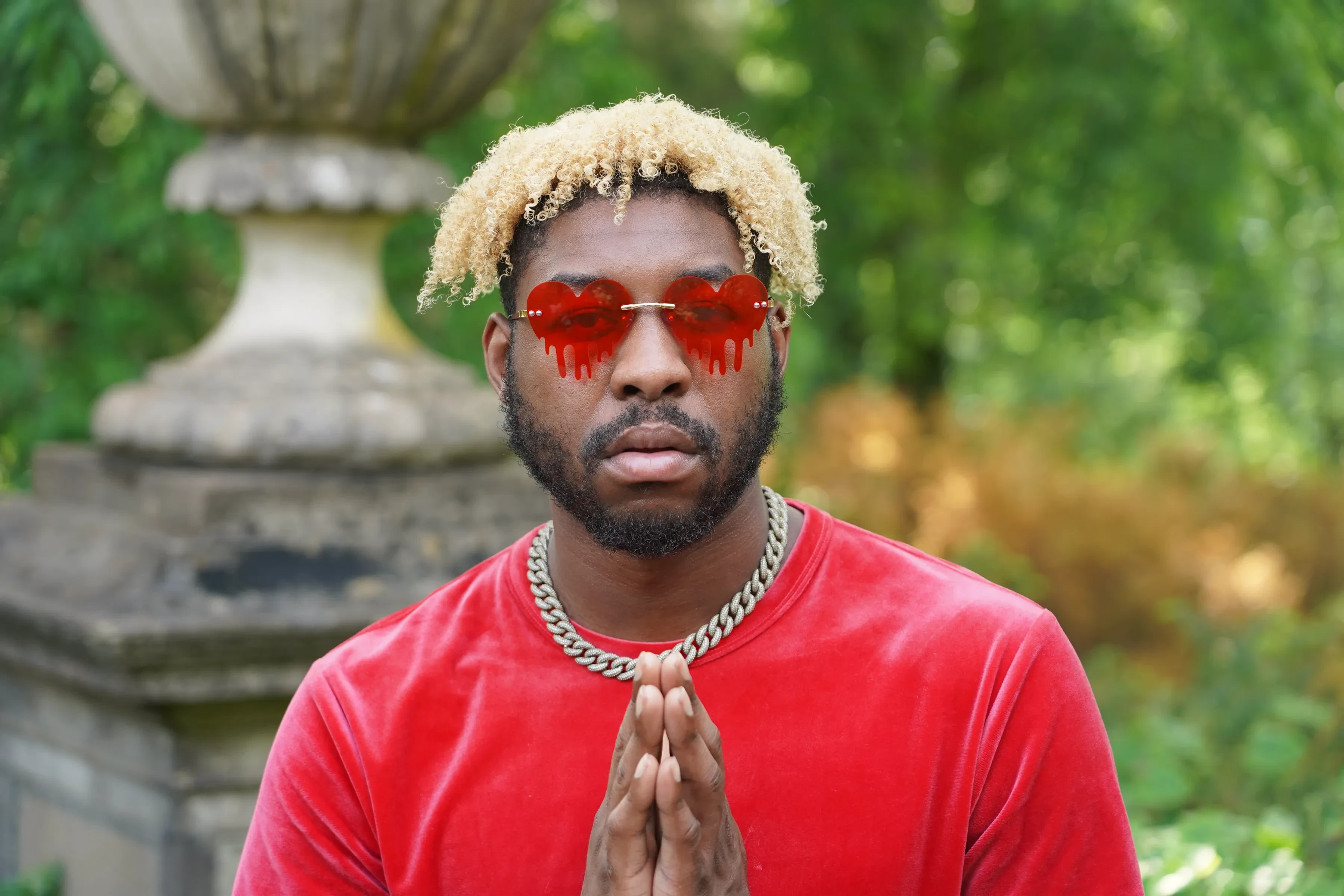Alpha The Wolf with both palms pressed together in front of his chest in a prayer gesture, looking straight down the lens through red heart lenses, a stone urn and green garden behind him