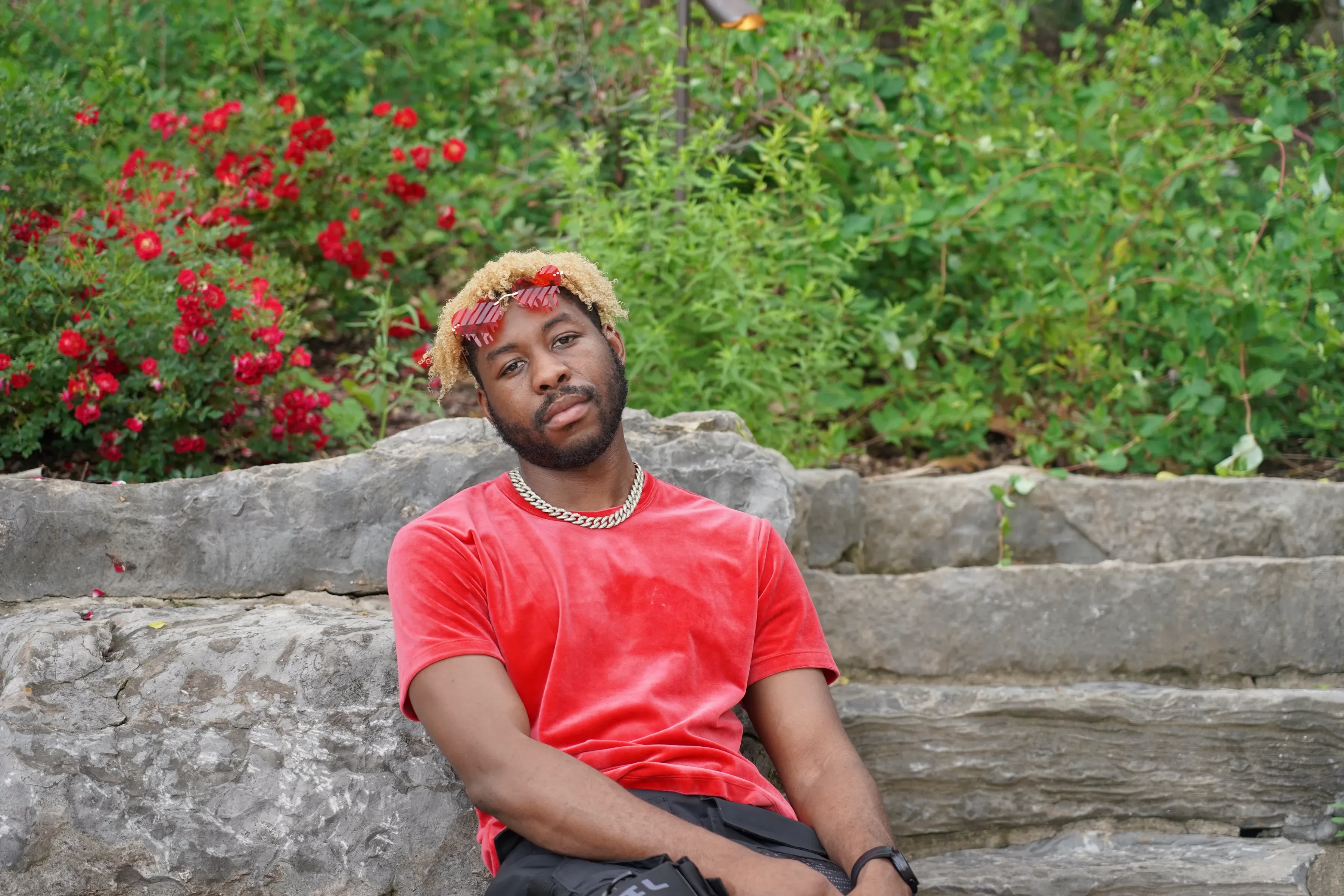 Alpha The Wolf seated on rough limestone block steps with his sunglasses pushed up into his hair, a bank of red roses in full bloom behind him
