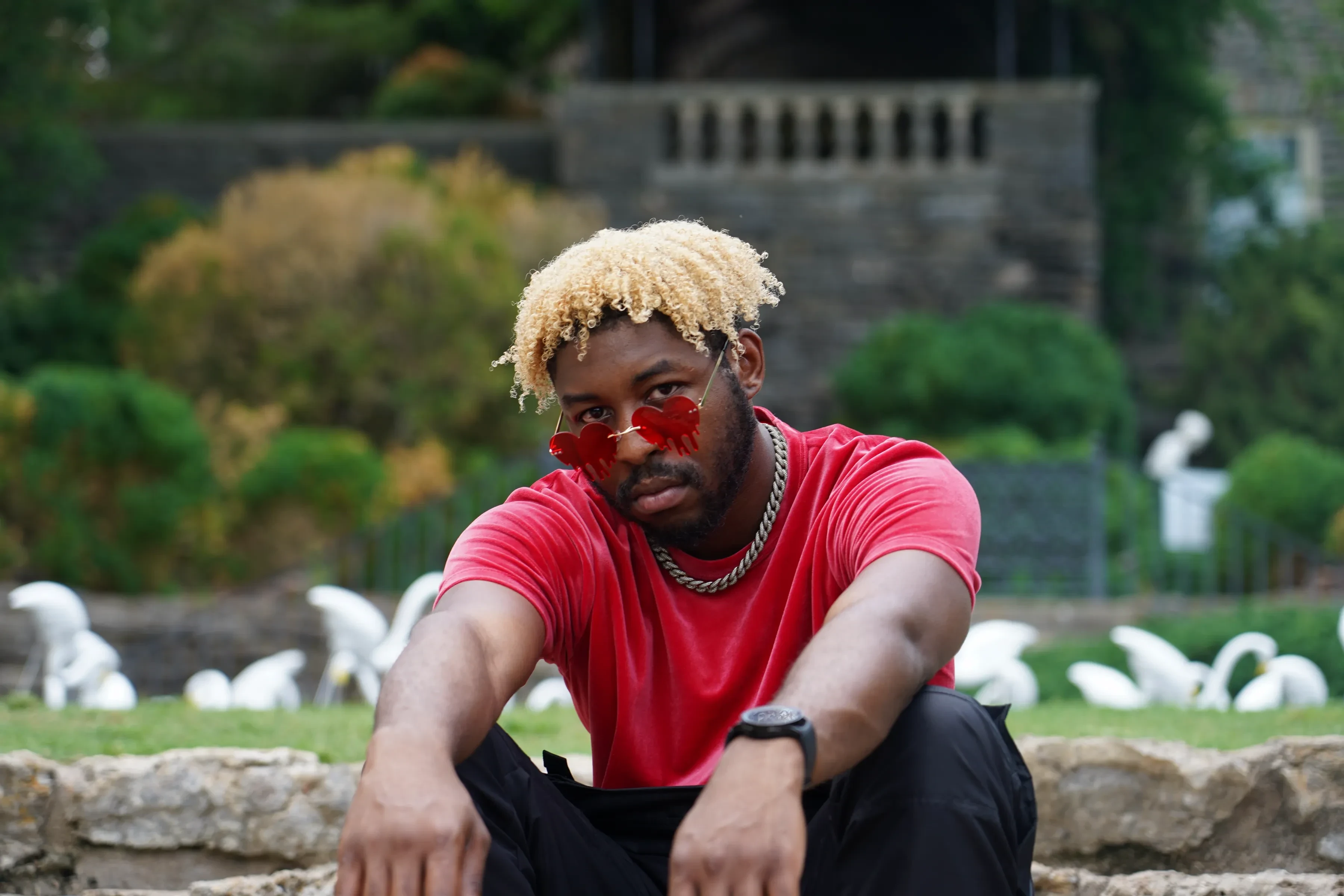 Alpha The Wolf sitting with his forearms draped over his knees, looking straight down the lens through red heart lenses, white flamingo lawn sculptures scattered across the grass behind him