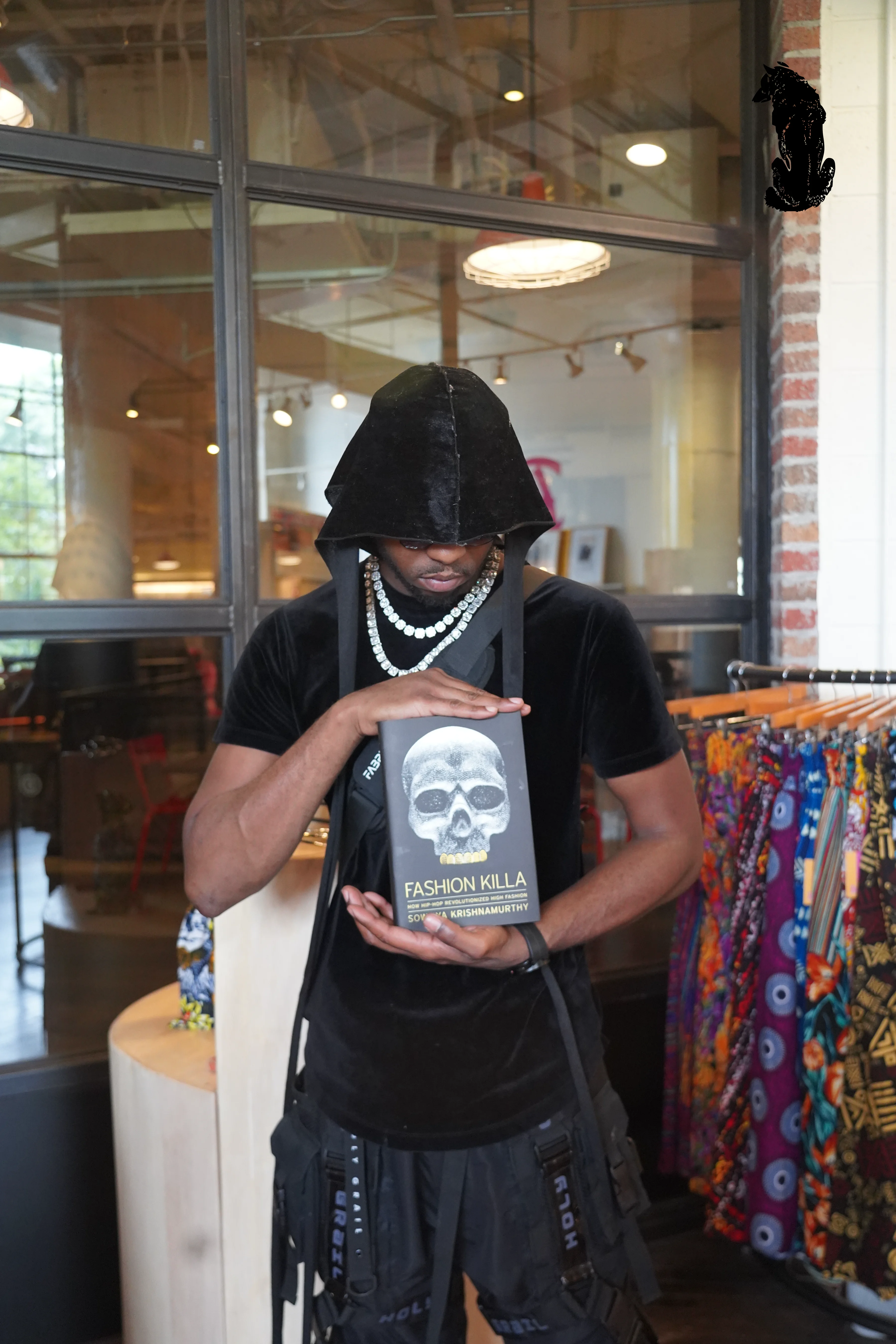 Alpha The Wolf inside a boutique, head bowed over a copy of 'Fashion Killa' by Sowmya Krishnamurthy which he holds up with both hands, draped in a black velvet hoodie with layered pearl necklaces and a crossbody bag.
