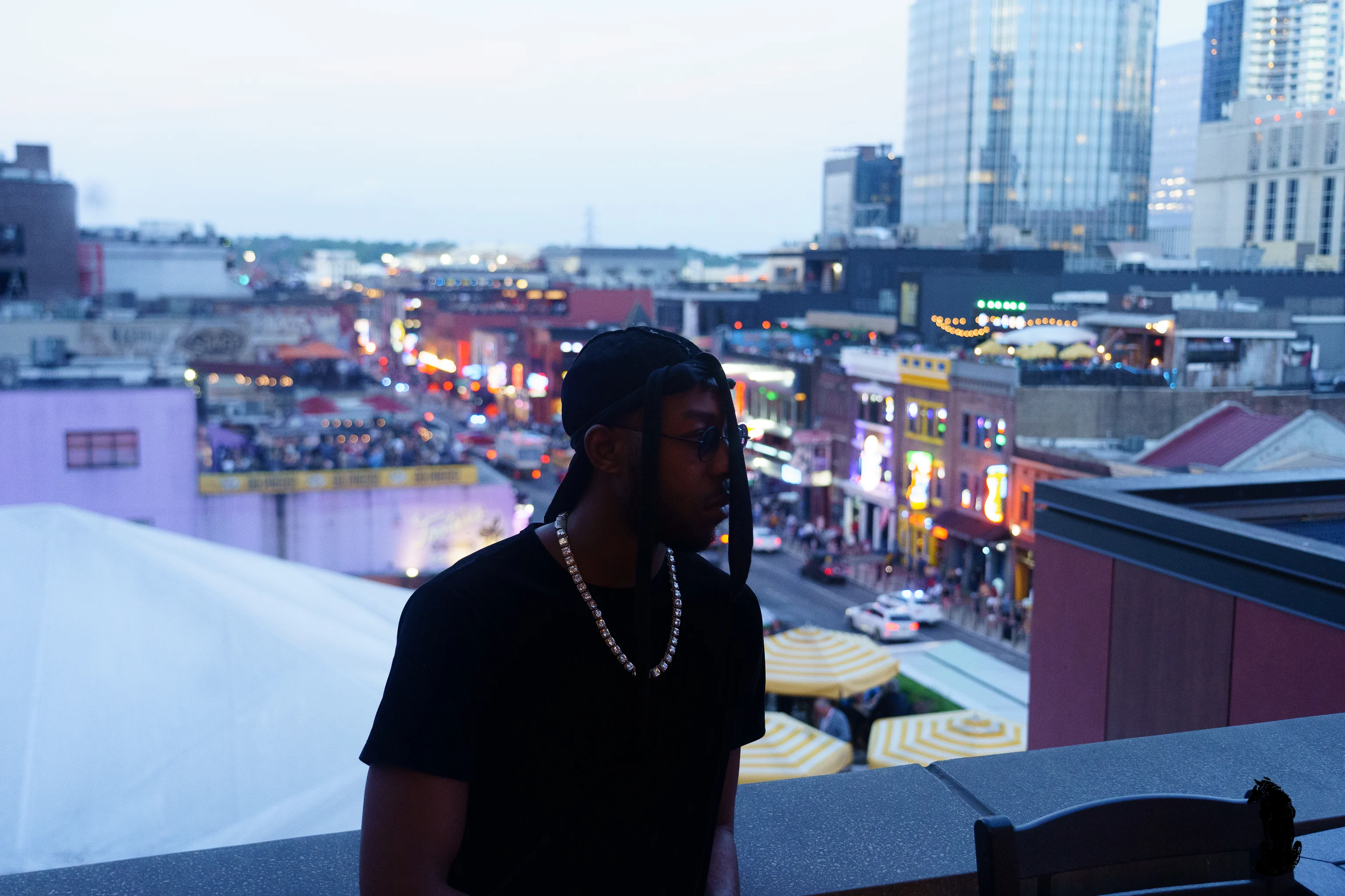 Alpha The Wolf in profile on a Nashville rooftop at blue hour, chin lifted, dark sunglasses on, the bokeh of Lower Broadway's neon signs bleeding into the backdrop behind him.