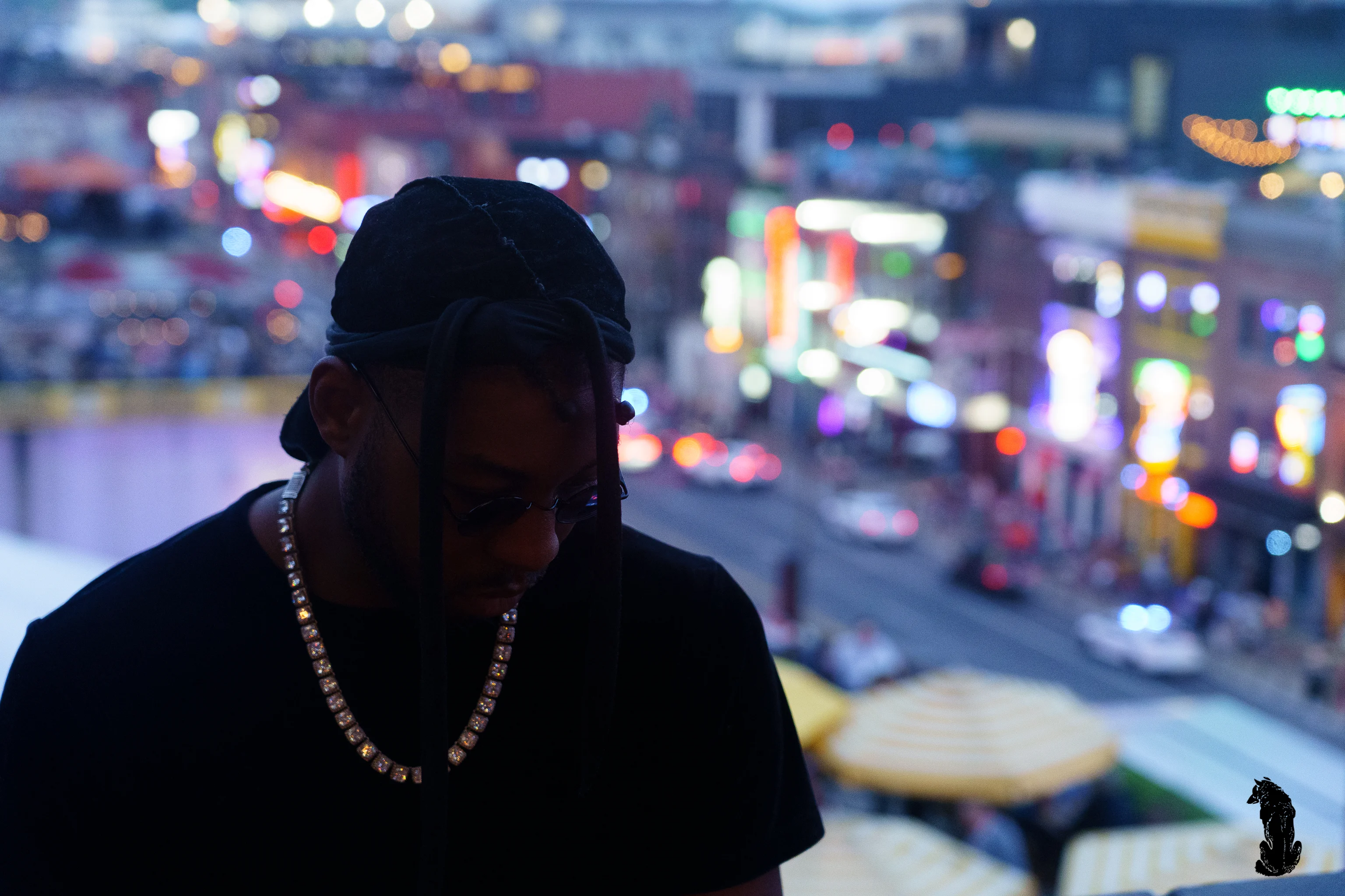 Alpha The Wolf in tight three-quarter profile, eyes hidden behind the brim of a black cap as the city lights of Nashville dissolve into colorful bokeh behind him.