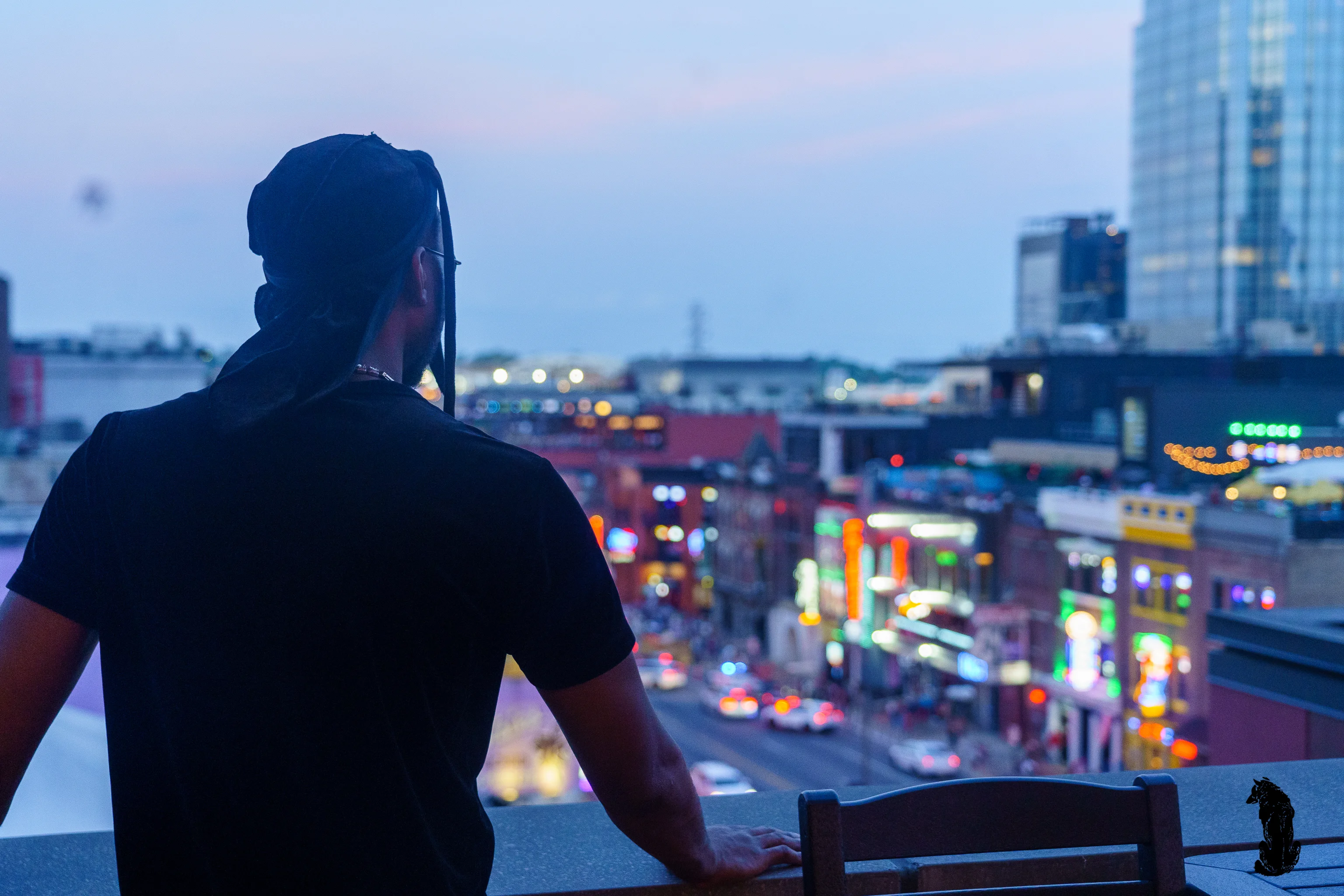 Alpha The Wolf shot from behind, standing at a rooftop railing and gazing out over Nashville's glowing Broadway strip at dusk, his silhouette framed against the wide cityscape