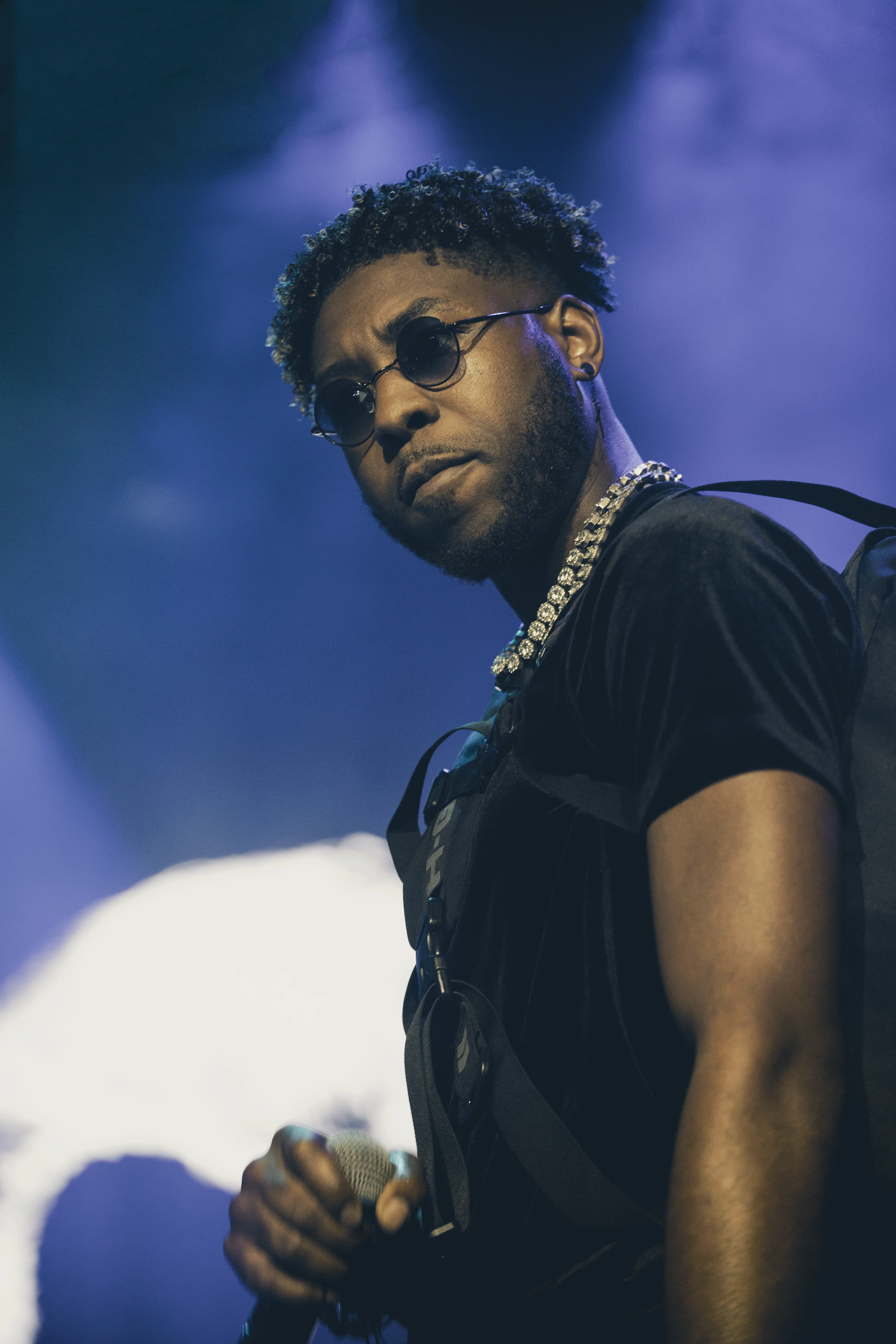 Alpha The Wolf on stage in a tight low-angle close-up, small round glasses and a chain catching the blue stage wash, microphone in hand and eyes cast slightly downward with focused intensity, the background dissolving into hazy violet-blue stage fog
