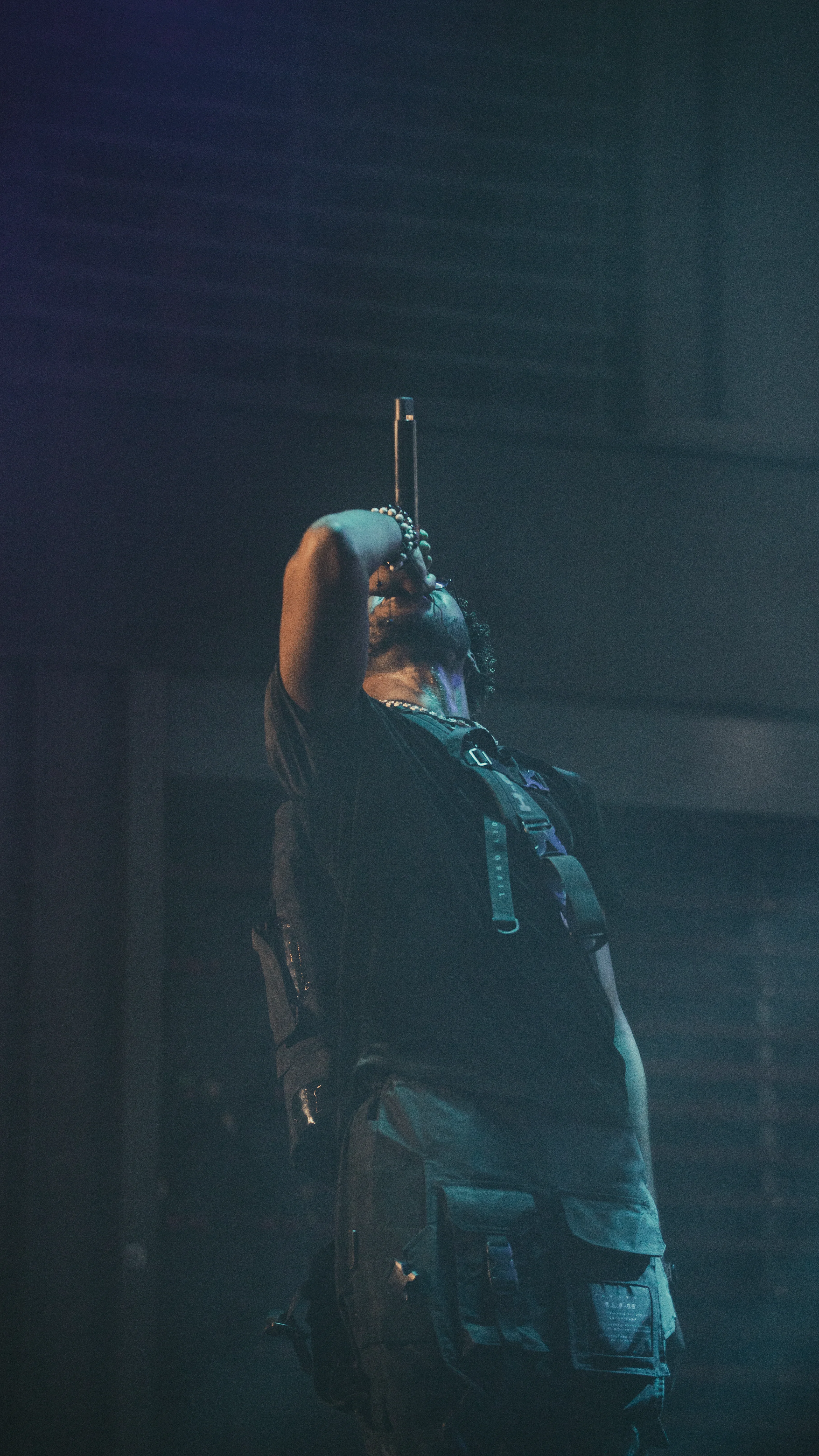 Alpha The Wolf shot from a distance, head thrown back and mic arm raised straight up toward the rigging, body arched in a dramatic release, the full-length silhouette swallowed by the dark stage with only teal-blue fog wrapping around him