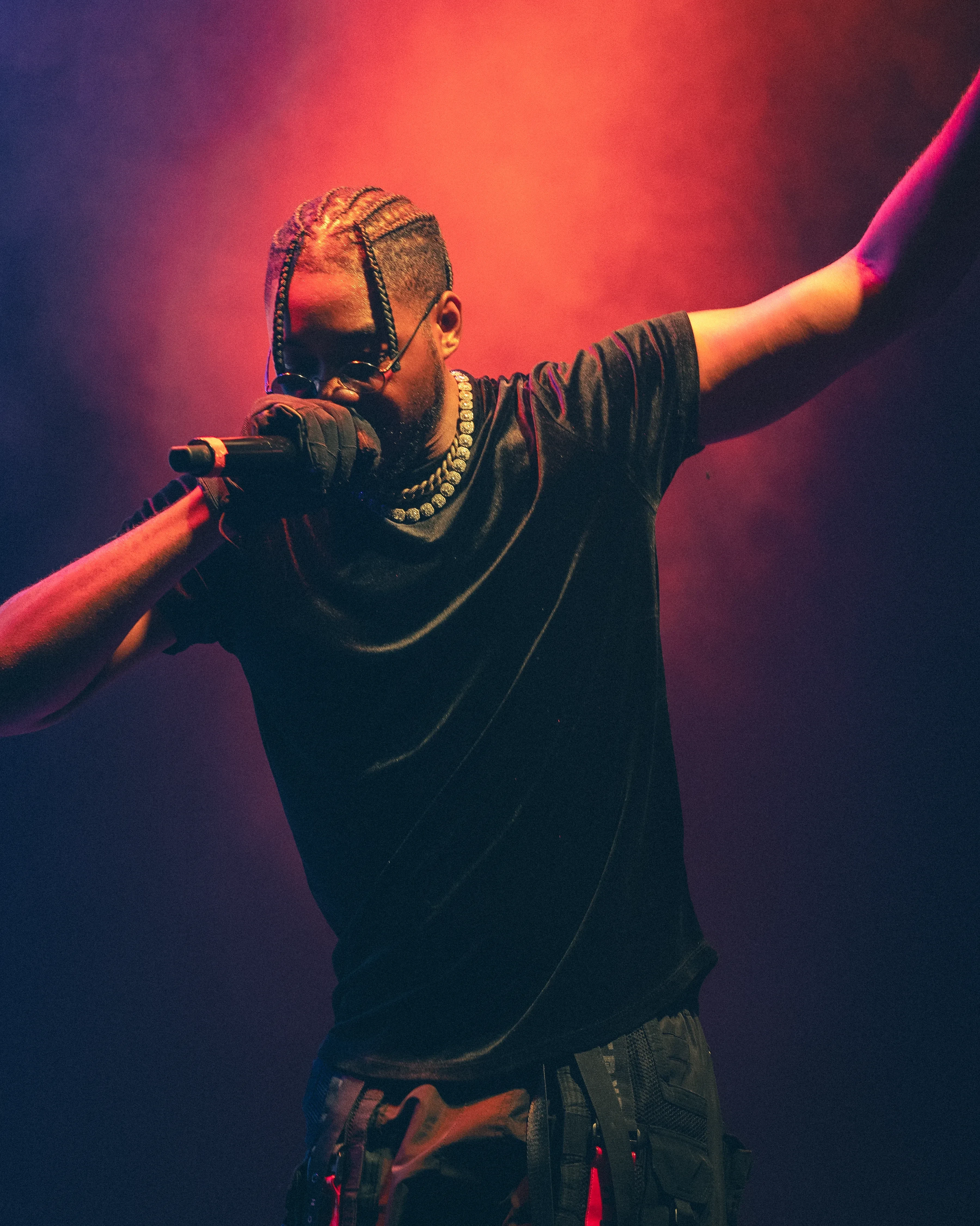 Alpha The Wolf with both arms flung out and the mic pressed to his mouth, black tee and strapped cargo pants, a red glow blooming behind his shoulders.