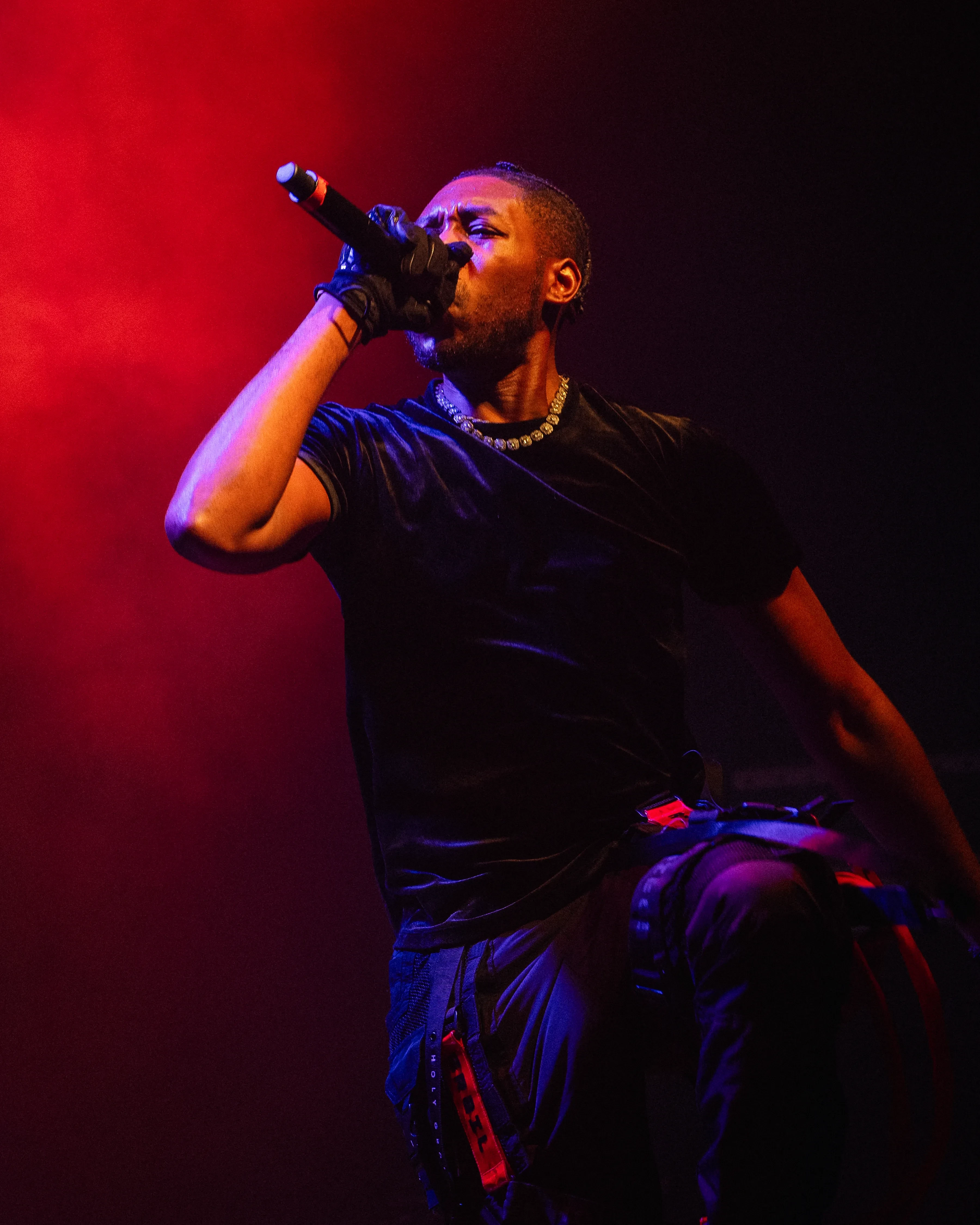 Alpha The Wolf standing with the mic raised and head turned, black tee and strapped cargo pants, red and purple light pooling in the smoke behind him.